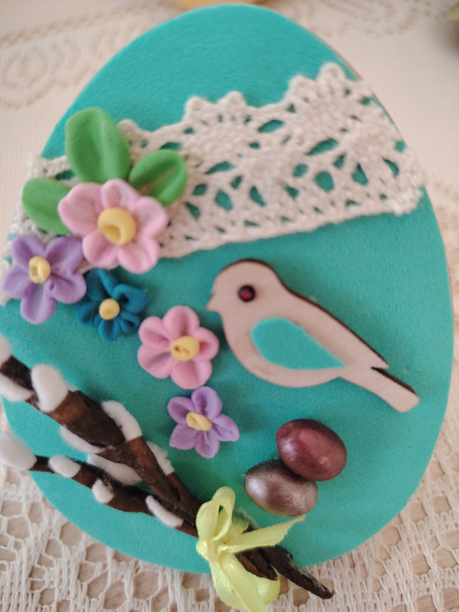 A close-up photograph shows an Easter egg decoration. The egg is teal-colored and has a white lace trim along the top edge. On the front of the egg are decorations including: a bird cutout, several small flowers of pink, purple, and blue with yellow centers, branches with pussy willow catkins, small egg-shaped objects, and a yellow ribbon. The egg is resting on a lacy white surface. The photo was taken in Sarapul, Russia.