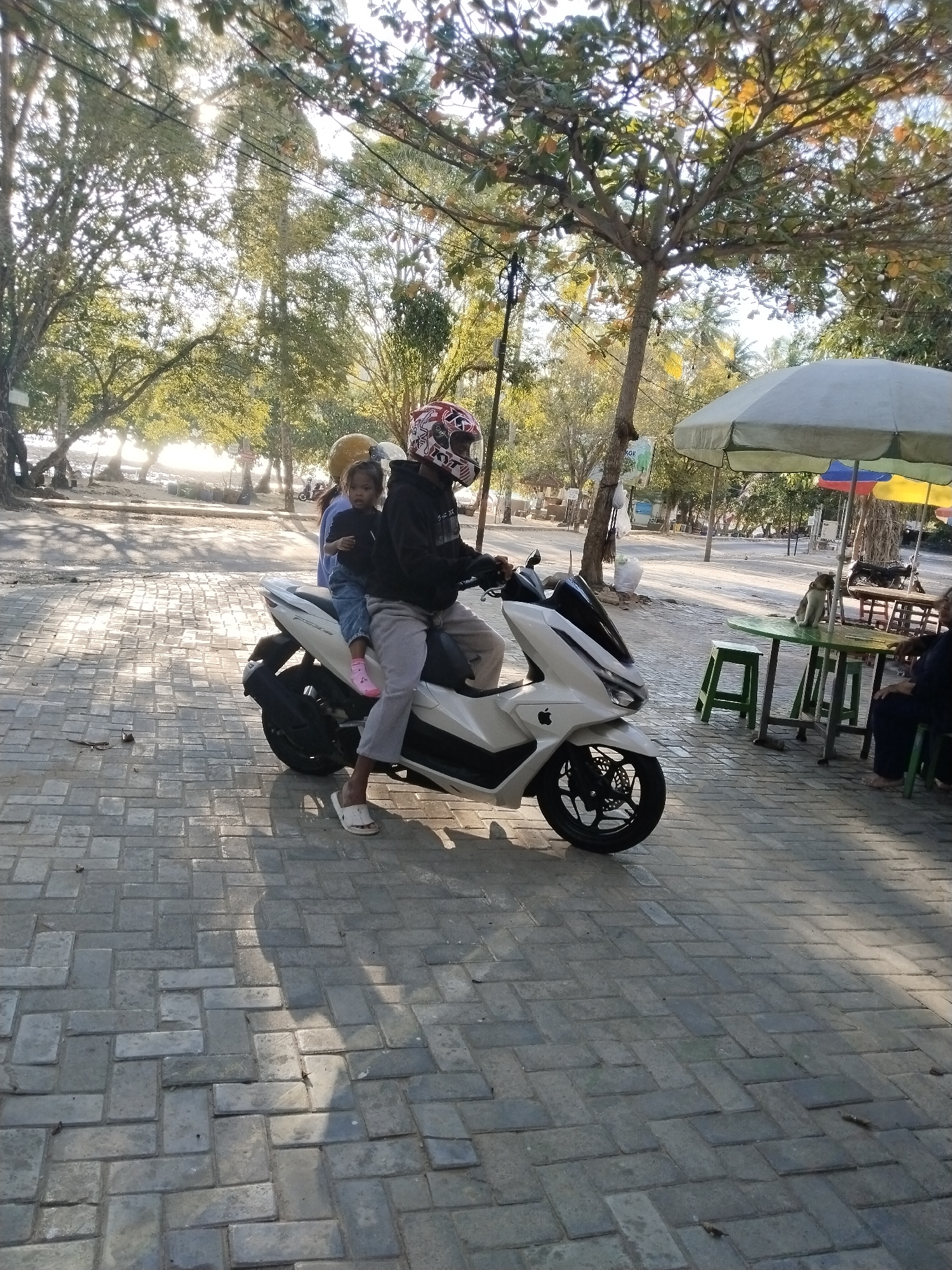 A man, a child, and a woman are on a white scooter. The man is driving, wearing a black hoodie and a red and white helmet. The child, wearing a blue shirt, sits between the man and the woman. The woman is in the back, wearing a yellow hat and a blue shirt. They are on a paved path in front of trees and a beach. In the background, there are tables and umbrellas, with a dog on one of the tables. This scene is located in Prigi, Indonesia.