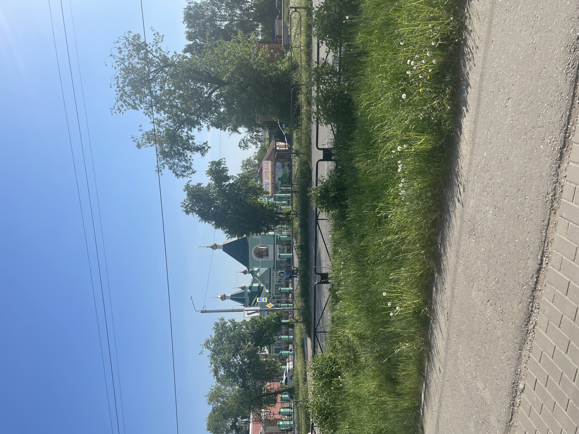 The image depicts a street scene in the city of Chaikovsky, Russia. A sidewalk runs along the right side of the image, with grass growing along its edge. A black metal fence separates the sidewalk from the road. Beyond the fence is a Russian Orthodox church with a light green facade and multiple golden domes. Trees are interspersed along the street, partially obscuring the church. In the background, other buildings and signage are visible. Power lines run across the blue sky. The image appears to have been taken during the day with bright sunlight.