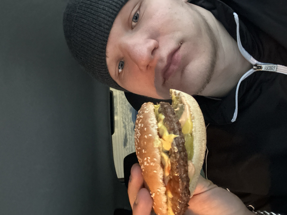 A male individual is positioned in the right foreground, partially obscuring the view of the background. He wears a dark grey knitted beanie and a black zip-up jacket with a white zipper, displaying a "NIKE" logo on the zipper pull. His blue eyes are visible, and he has light facial hair and a potential lip piercing on the lower right side of his mouth. He is holding a double cheeseburger in his left hand, presenting it towards the viewer. The burger consists of a sesame seed bun, two beef patties, melted yellow cheese, sliced pickles, and an orange/pinkish sauce. The bottom bun shows an irregular edge, possibly from a bite. The background suggests the interior of a vehicle, with a dark headliner and a rear window showing a dimly lit exterior with faint horizontal reflections, indicative of a night or dusk setting with potential snow or ice outside. The precise location within Div'ya, Russia, is not discernible from the image content.
