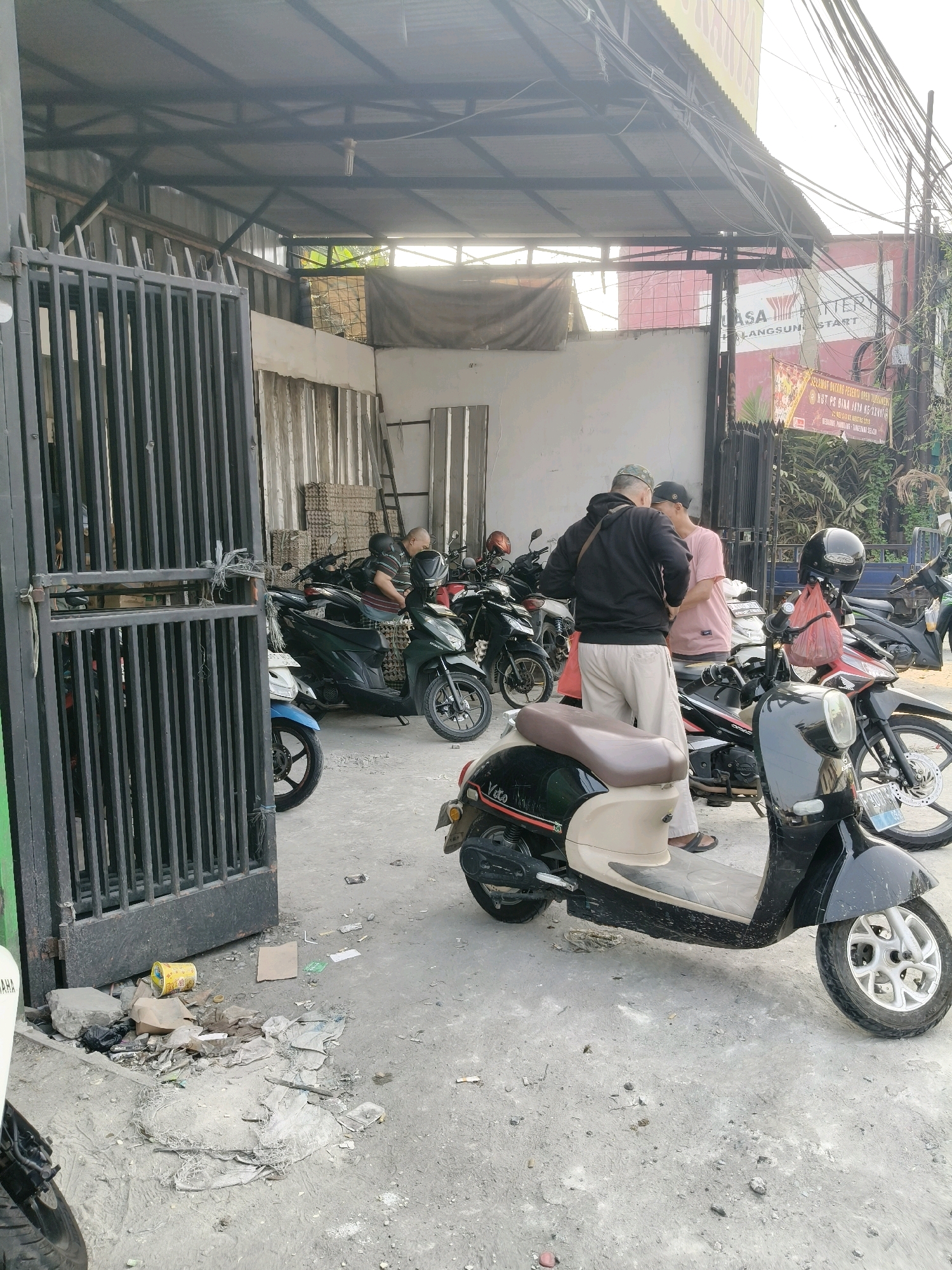 Two men are standing and talking outside a covered motorcycle repair shop in South Tangerang, Indonesia. One man, wearing a black hoodie and light-colored pants, is facing away from the camera. The other man, in a pink t-shirt and black cap, is facing the first man. Several motorcycles are parked in the shop, some with helmets and bags attached. In front of the shop, a black and cream colored scooter is parked. The ground is paved with concrete and is littered with debris, including plastic bags, paper, and a yellow can. A metal gate is partially open to the left of the scene. The background shows corrugated metal walls, electrical wires, and a sign with red lettering. The sky is bright and hazy.
