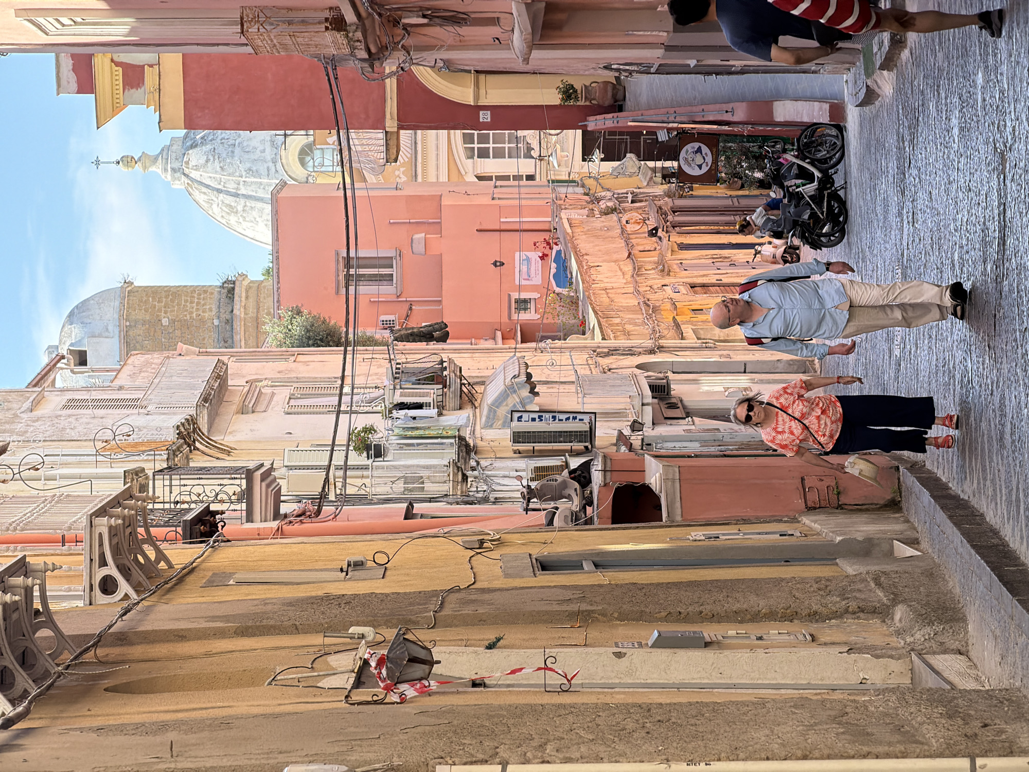 This image depicts a narrow street in Procida, Italy. Buildings of various colors including yellow, peach, and pink line the street on both sides. Two people, a man and a woman, are walking on the right side of the street. The woman is wearing a red and white patterned shirt and dark pants, while the man is wearing a light blue shirt and khaki pants. Behind them are other pedestrians, and parked bicycles and scooters. Above the buildings, a dome can be seen against a blue sky. Wires and cables crisscross above the street. Air conditioning units and satellite dishes are attached to the buildings.