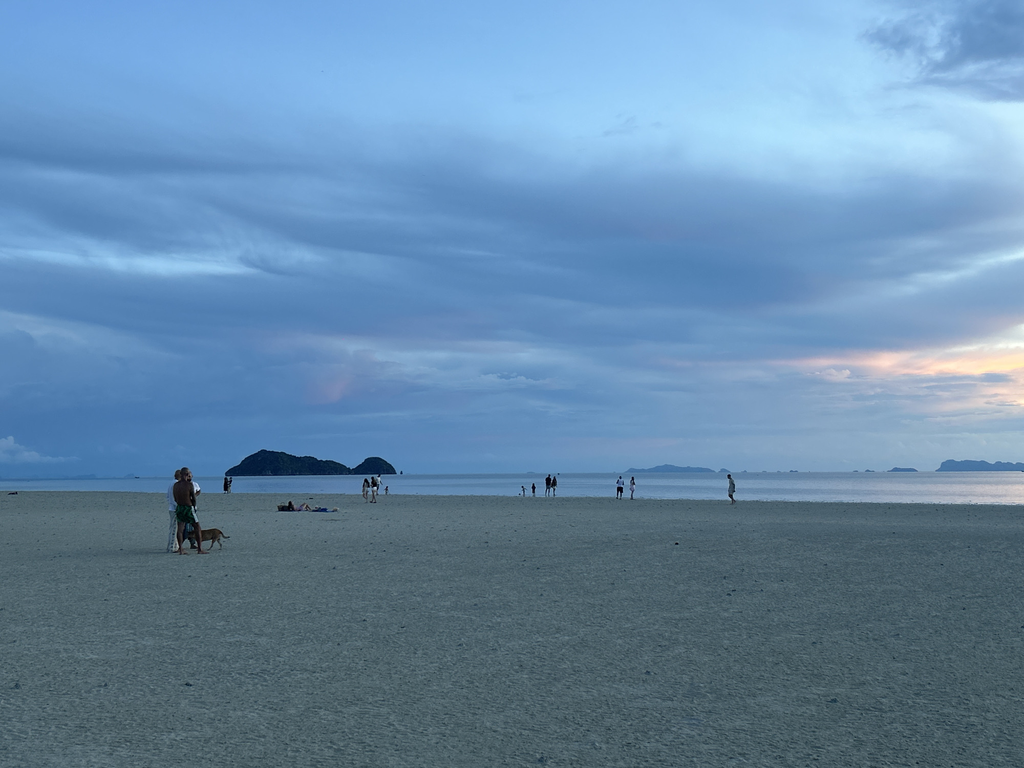 A wide, sandy beach on Ko Pha Ngan, Thailand, is captured at dusk. The sky is a gradient of blues, with wispy, darker clouds scattered across the horizon, and a faint glow of orange and pink light is visible where the sun has recently set. The sea stretches out to meet the sky, calm and reflective. In the distance, several small, dark islands dot the water. On the beach, a few people are spread out, engaged in various activities. In the foreground, a man in green shorts and a white shirt stands with another person, and a small dog is at their feet. Further down the beach, other individuals are walking, standing, or relaxing on the sand. The overall scene is tranquil, with the muted light of twilight creating a serene atmosphere.