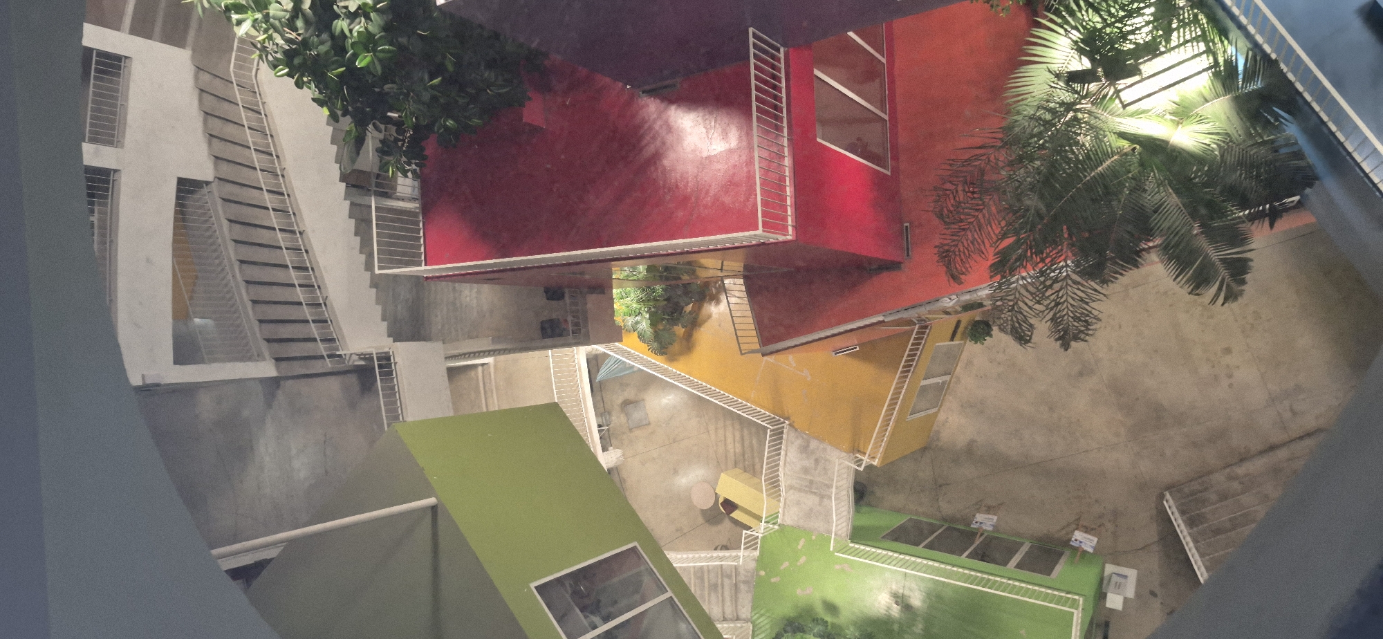 The image is an aerial view of a multi-level outdoor structure with brightly colored buildings and stairs. The structure appears to be modern and designed with intersecting planes and vibrant hues.
The dominant colors are green, yellow, and red, which form the facades of the buildings or platforms. White railings and staircases are interspersed throughout, providing access between different levels. There are several outdoor staircases, some of which appear to be expansive and lead to upper levels. Plants, including trees with large fronds, are visible in some areas, adding a touch of greenery and softness to the geometric architecture.
The ground level and walkways are paved with concrete or a similar material. The lighting suggests it is either evening or nighttime, with artificial lights illuminating some areas and casting shadows. There are no people visible in the image.
This structure resembles part of the pyramid and surrounding complex located in Tirana, Albania. The original pyramid building, now a cultural center and former museum, has undergone renovations, and this scene could depict a section of its modernized exterior, particularly the areas that have been redeveloped with new architectural elements and vibrant colors.