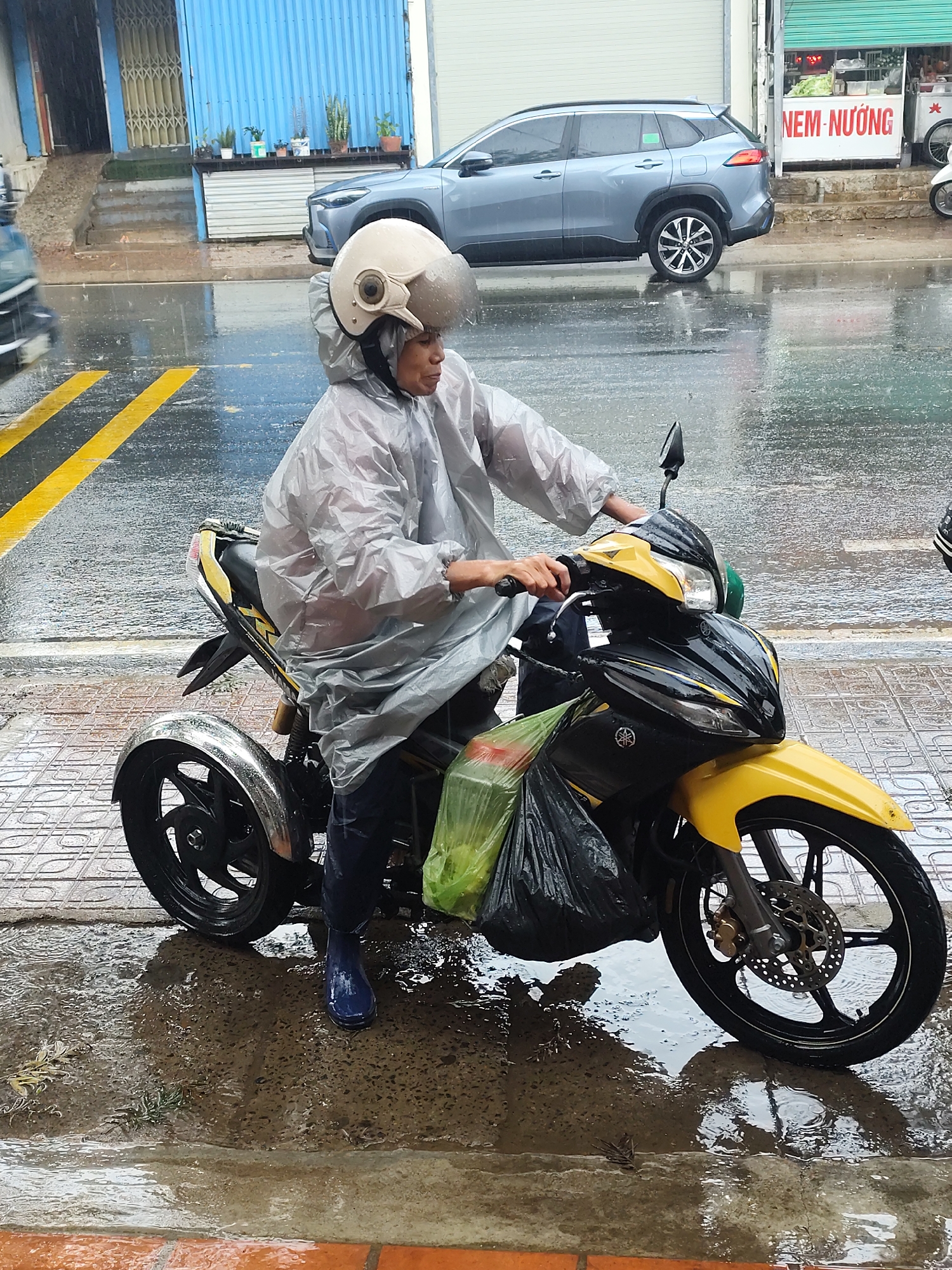 A person wearing a cream-colored helmet and a clear poncho is sitting on a yellow and black Yamaha motorcycle on a wet, tiled sidewalk in what appears to be a rainy urban setting. The person is also wearing blue rubber boots. Two plastic bags, one black and one green, hang from the motorcycle.
In the background, there is a light blue building with a metal roll-up door and several potted plants on a ledge in front of it. A light blue SUV is parked on the wet street in front of the building. There is a sign written in Vietnamese above a food cart or small business on the right side of the frame, with the words "NEM NƯỚNG" visible. Yellow lane markings are visible on the street, and the general environment suggests a city, specifically Lang Biang - Đà Lạt, Vietnam, due to the signage and architectural style.