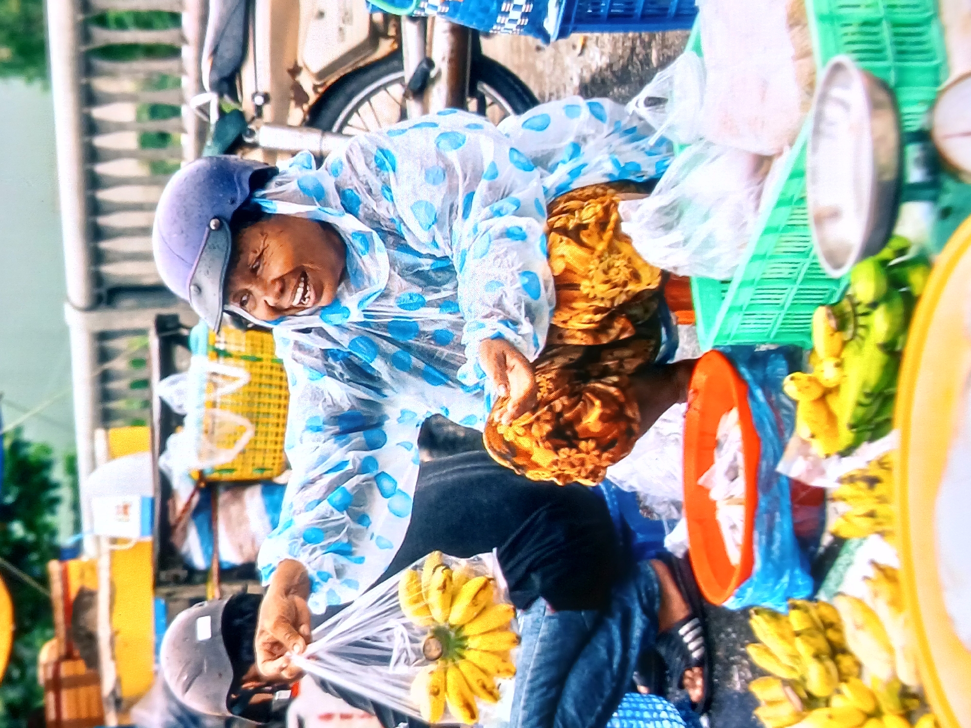 A woman in a blue polka dot poncho laughs while selling bananas from a street stall. She is wearing a blue helmet and a patterned skirt. In front of her, several bunches of bananas are displayed on a blue tarp. Behind her, a motorcycle and other market stalls are visible. The scene is set in a busy urban environment.