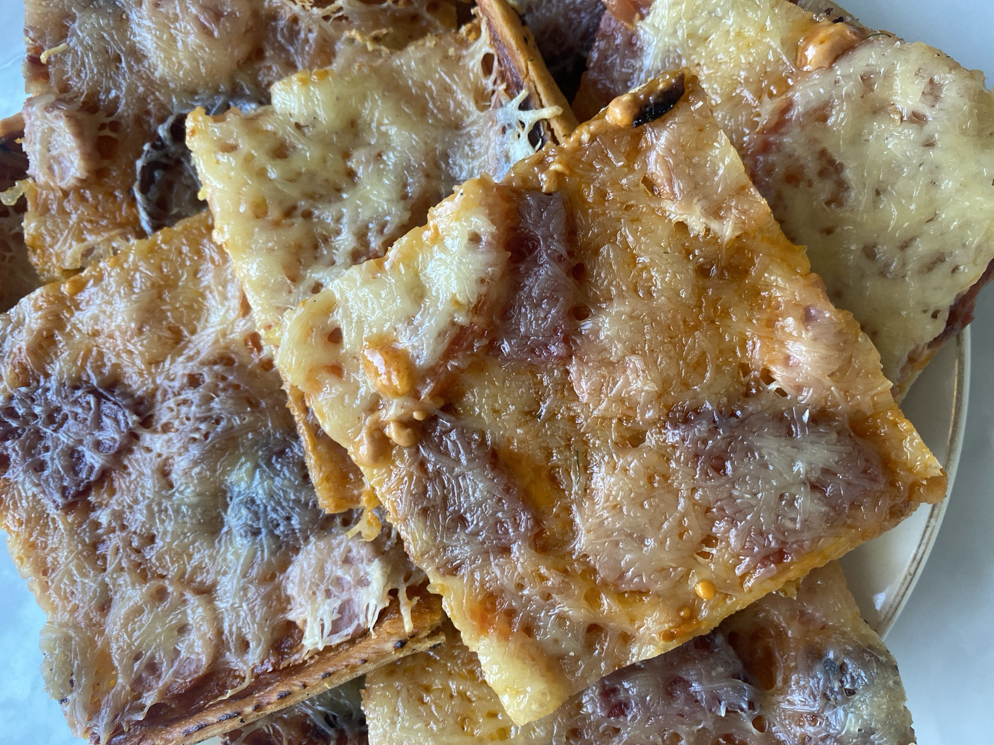 A close-up, overhead shot shows a pile of sliced pizza on a plate. The pizza has a thin crust, a red sauce, and a generous amount of melted cheese that has browned in some areas. The slices are irregular in shape and overlap each other, making it difficult to discern the exact number of pieces. The plate has a thin, decorative rim. The image is sharp and well-lit, focusing on the texture and appearance of the pizza. The location in the city of Mamontovo, Russia, is not depicted in the image.