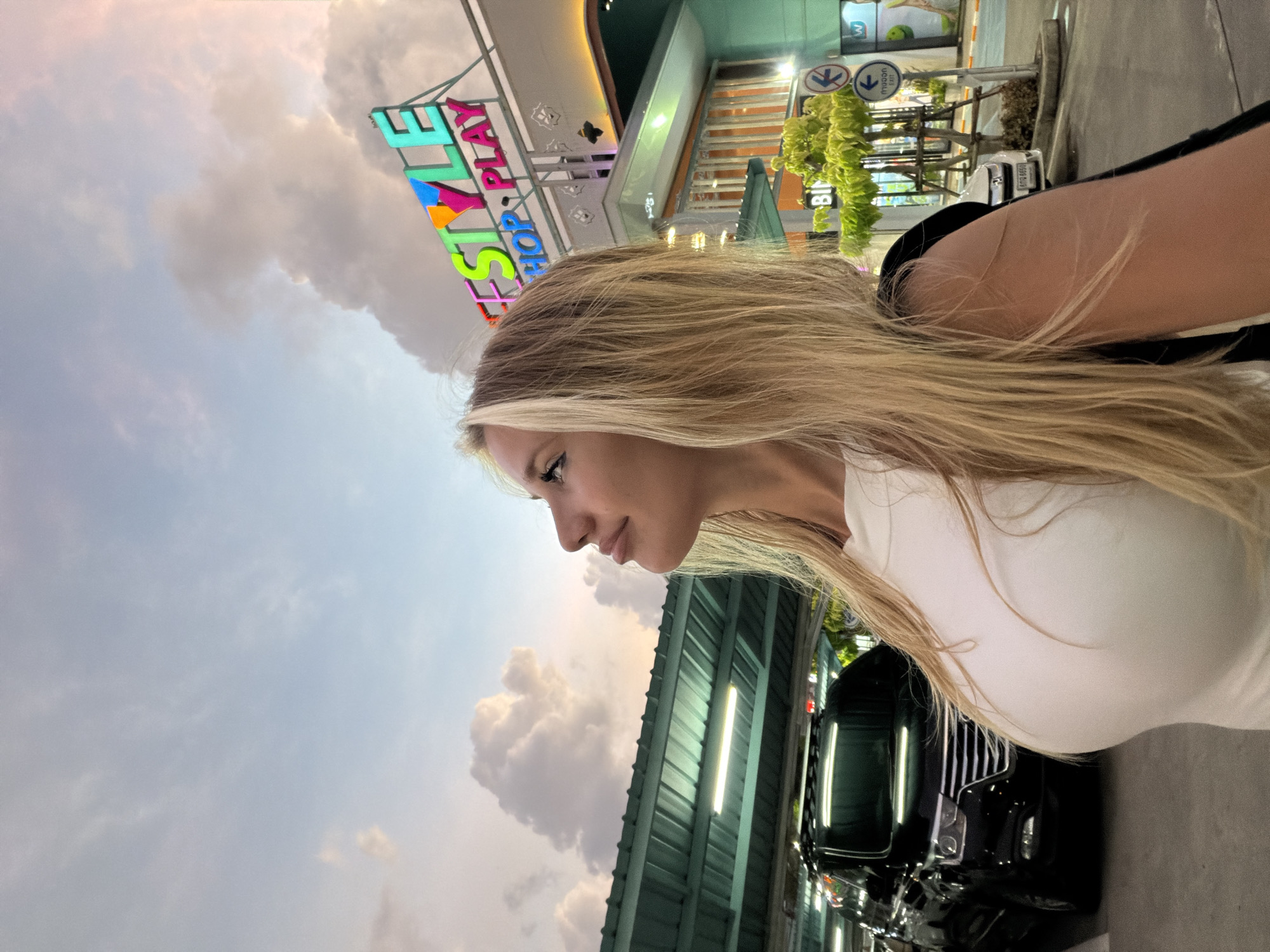 A blonde woman is posing for a photograph in an outdoor parking lot in Si Sunthon, Thailand, during twilight. She is looking upwards and to her left, with her head tilted back. The woman is wearing a white sleeveless top and a black jacket draped over her shoulders. Behind her, a black van is parked under a covered structure with green beams and fluorescent lights. Further in the background, a building with colorful neon signage that reads "LIFESTYLE SHOP PLAY" is visible. The sky displays a mix of soft pink and blue hues with fluffy clouds, indicating the transition from day to night.