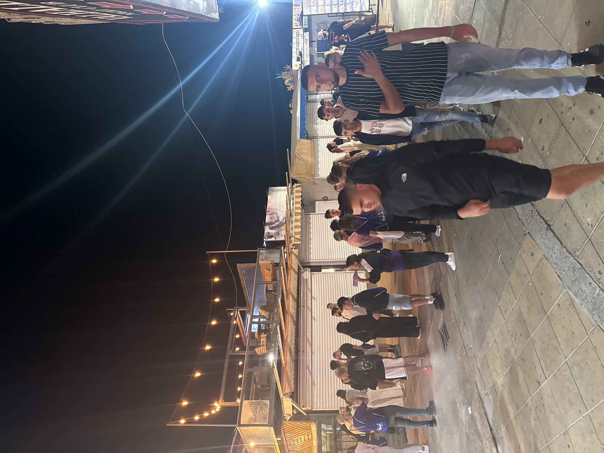 The image depicts a street scene at night in Nesebar, Bulgaria, likely a tourist area due to the presence of illuminated string lights and various stalls or shops. A group of young people are walking and standing on a tiled walkway. In the foreground, two young men are walking towards the camera. The man on the right is wearing a black and white striped t-shirt and light blue jeans, with his right hand on his chest. The man on the left is wearing a black hooded jacket and black shorts. Behind them, a diverse group of individuals, both male and female, are seen interacting and walking. The background features a multi-story structure with open-air seating and shops with closed shutters, suggesting it may be closing time or off-season. The overall atmosphere appears lively, with artificial lighting illuminating the area.