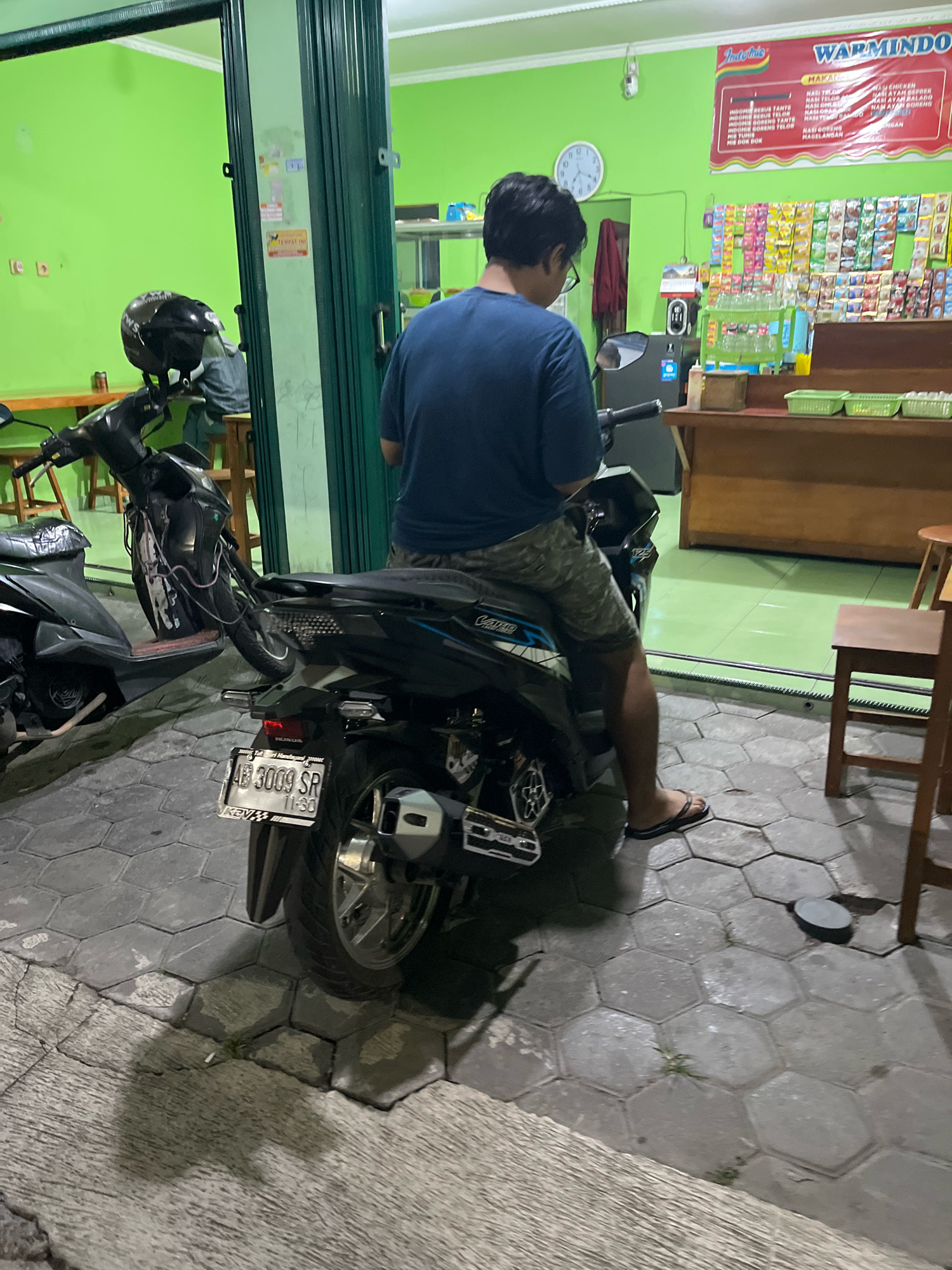 In Banguntapan, Indonesia, a person wearing a blue t-shirt and camouflage shorts is sitting on a black motorcycle, facing away from the camera. The motorcycle has a license plate reading "AB 3009 SR 11-30." In the background, a bright green wall is visible, along with a counter displaying various products and a menu board. To the left, another motorcycle with a black helmet on its handlebars is parked. The ground is paved with hexagonal concrete tiles.