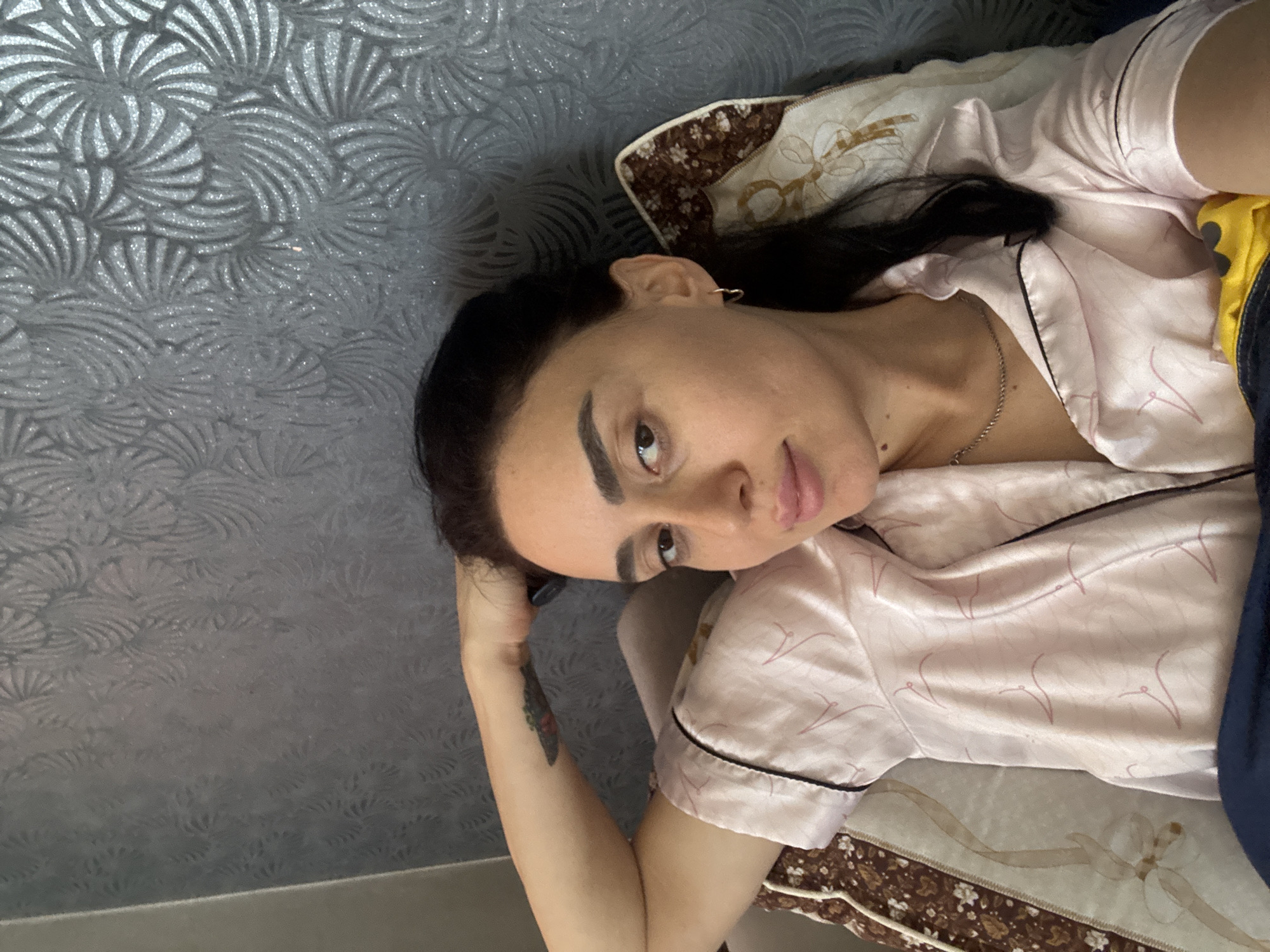 A woman is lying on a bed. She has dark hair and is wearing a pink pajama top with black piping. She is looking at the camera. The background is a patterned wall. The image was taken indoors. There is a pillow behind her head. Her arm is bent. There is a tattoo visible on her forearm. Her location in the city of Tbilisi, Georgia, cannot be determined from the image.