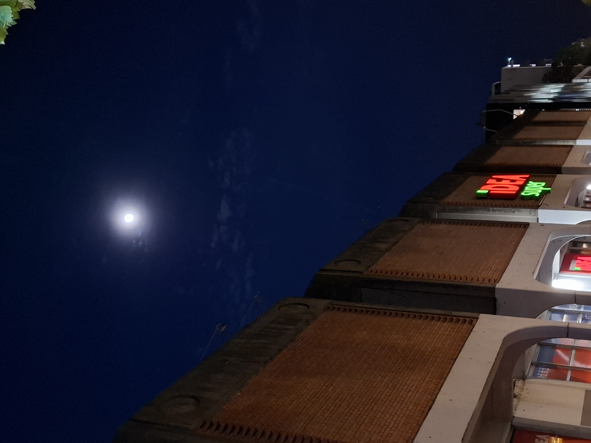 This nighttime photograph captures an urban scene with a dominant dark blue sky and a commercial building. The image is taken from a low angle, tilting upwards and to the right, presenting a large expanse of the deep blue night sky on the left and top, contrasted with architectural structures on the right and bottom.
In the upper left quadrant of the sky, a bright, full or near-full moon glows prominently, surrounded by subtle, wispy cloud formations. The sky transitions from a deep indigo at the top to a slightly lighter blue closer to the horizon.
On the right side, a multi-story building features a distinctive facade. It appears to be constructed with sections of reddish-brown brickwork, arranged in a textured pattern, interspersed with lighter, possibly concrete or stone, architectural elements that include curved or arched structures. Near the top right of the building, a brightly illuminated neon sign is visible, displaying the word "IDEA" in red letters and "Super" in green letters, indicating a supermarket or large retail store. Other subtle lights from the building's interior or exterior are also visible. A small amount of green foliage, possibly from a tree or plant, can be seen in the extreme top-left corner of the frame.
The overall scene depicts a tranquil night in an urban environment, where the natural light of the moon coexists with the artificial illumination of city architecture. No specific activity or people are visible. The presence of the "IDEA Super" sign strongly suggests the location is consistent with Novi Sad, Serbia, where this supermarket chain operates.