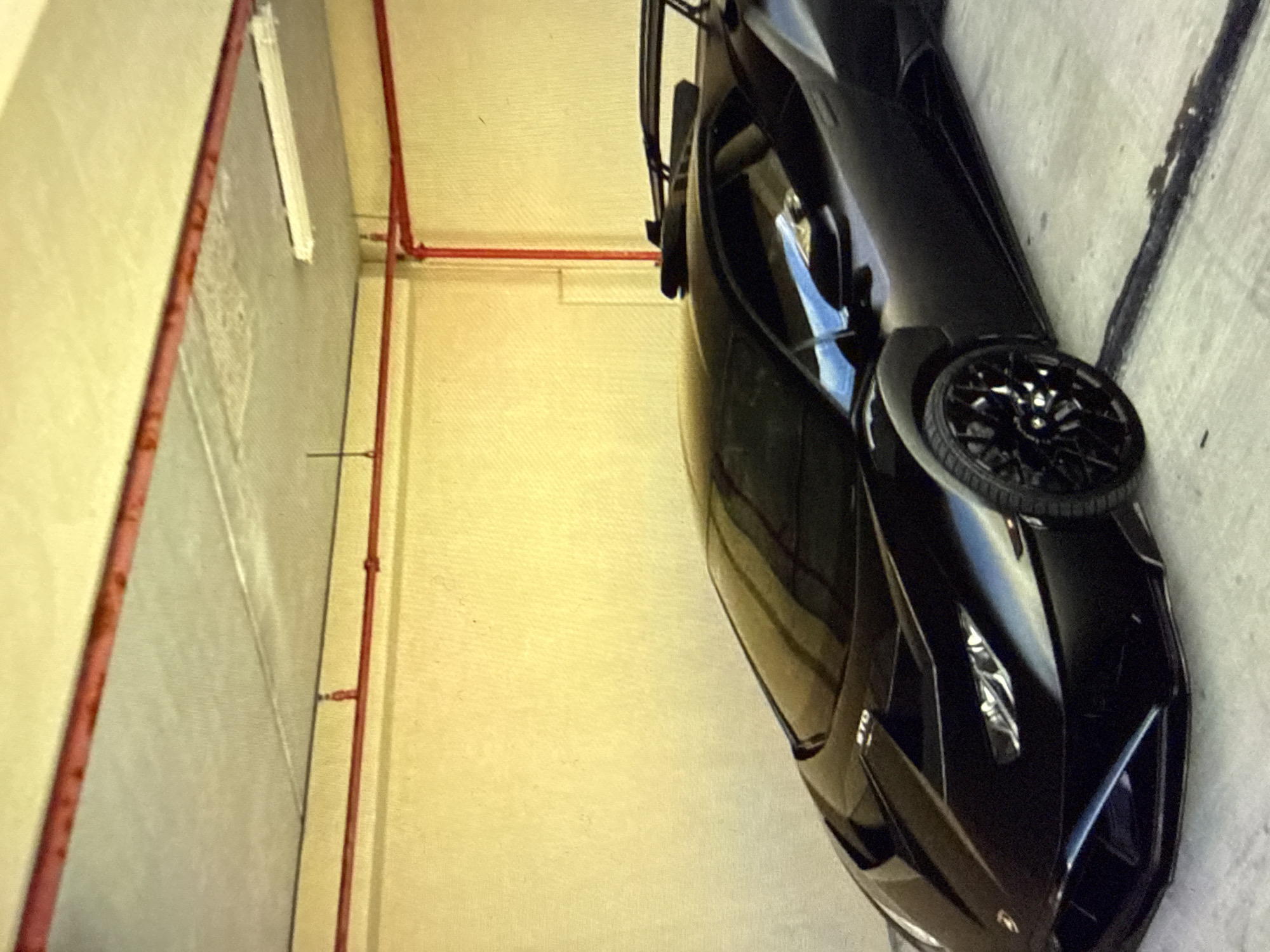 A black Lamborghini Huracán STO supercar is parked indoors in a fixed position, angled slightly towards the left of the frame. The vehicle is black with multi-spoke black wheels and features a prominent rear wing and aerodynamic front fascia. The letters "STO" are visible on the front bonnet. The surrounding environment consists of light yellow-beige textured walls and a grey concrete ceiling. A system of red pipes, consistent with a fire suppression system, runs along the upper walls and ceiling, with one sprinkler head visible. A single white fluorescent light fixture is mounted on the ceiling in the upper right. The concrete floor exhibits several dark, irregular stains, including a long, dark streak running parallel to the car's right side. No individuals are present. The location within the city of Kardzhali, Bulgaria, cannot be determined from the visual evidence provided.