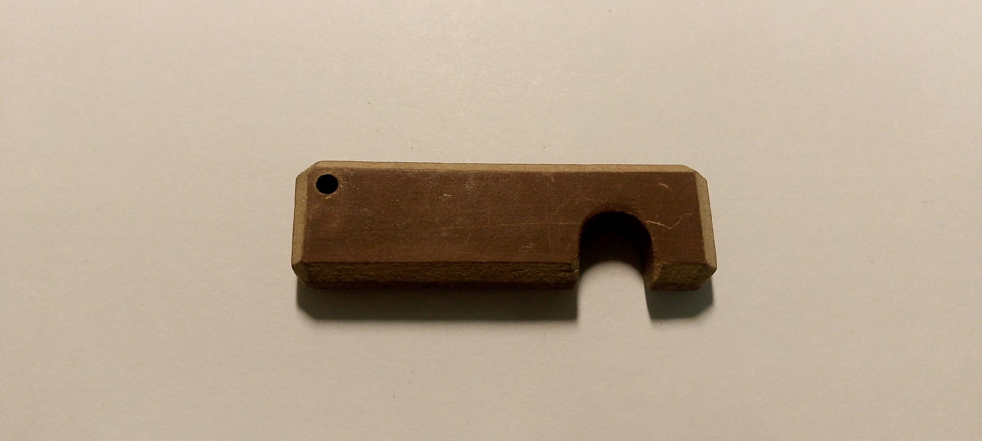The image shows a single, small, brown, rectangular object with a cutout on the right side and a small hole on the left side. The object appears to be made of wood or a similar material and is lying on a light-colored, flat surface, likely a table or floor. There are no people or any other objects visible in the image. The lighting is diffused, creating a soft shadow to the right and bottom of the object. There is no indication of the location in Zarechny, Russia, or any environmental context beyond the flat surface.