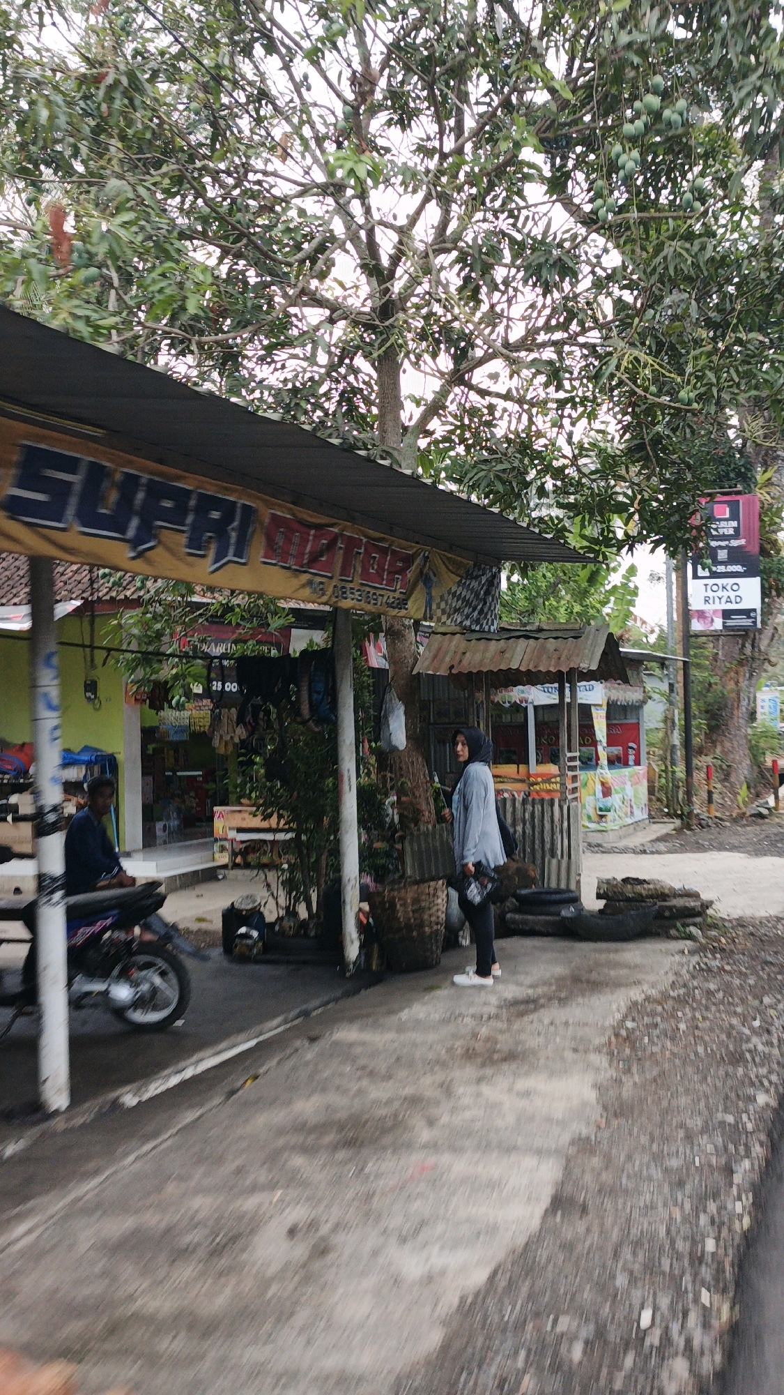 A person wearing a dark hijab and a light blue jacket stands to the right of a tree, holding a black bag. They are wearing white shoes. In the background, there is a stall with colorful offerings, and a large tree with green fruit hangs over a sign that reads "SUPRI MOTOR". To the left, another person sits on a motorcycle. The scene appears to be a roadside establishment in Sukaresik, Indonesia, with a paved walkway in the foreground and a dirt shoulder with scattered debris on the right.