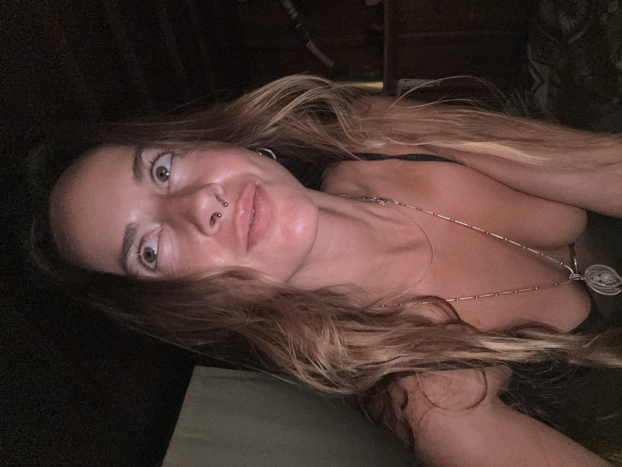 A medium shot shows a woman looking upwards and to the right, her head tilted slightly back. She has long, wavy blonde hair that falls around her shoulders. Her eyes are open and her eyebrows are raised. She has a septum piercing and a small hoop earring in her left ear. She is wearing a dark tank top and a long silver necklace with a circular pendant. The lighting is dim, with a warm glow highlighting her face. The background is dark and indistinct, suggesting an indoor setting at night.