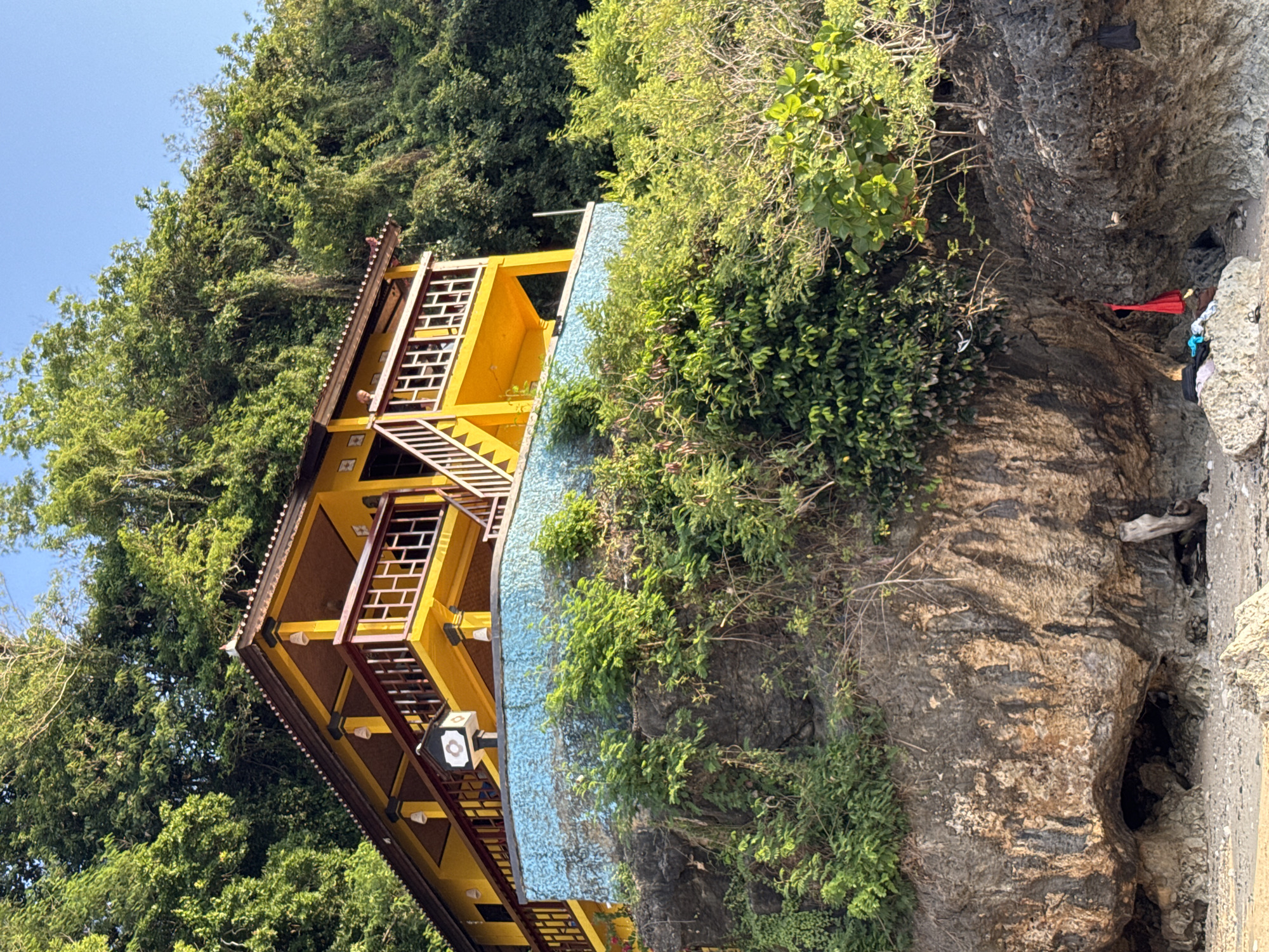 A sideways view shows a yellow, multi-story building perched on a rocky hillside in Kuta Selatan, Indonesia. The building features lattice-work railings and a blue tile-like exterior element. Lush green vegetation surrounds the building and extends up the hillside. The rock face is textured and partially covered in plants. In the rocky area at the base, there are scattered items, including what appear to be pieces of cloth. The sky is a clear blue.