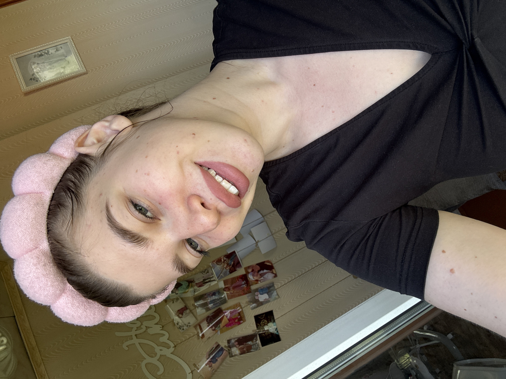 A woman with a pink headband is posing for a selfie. She is wearing a black top with a knot detail at the neckline. Her skin is fair with some visible blemishes. She is smiling slightly, showing her teeth. The background includes a wall with a framed picture and several smaller images taped to it. The scene appears to be indoors, near a window with a glimpse of an outdoor setting. The location is not discernible from the image.