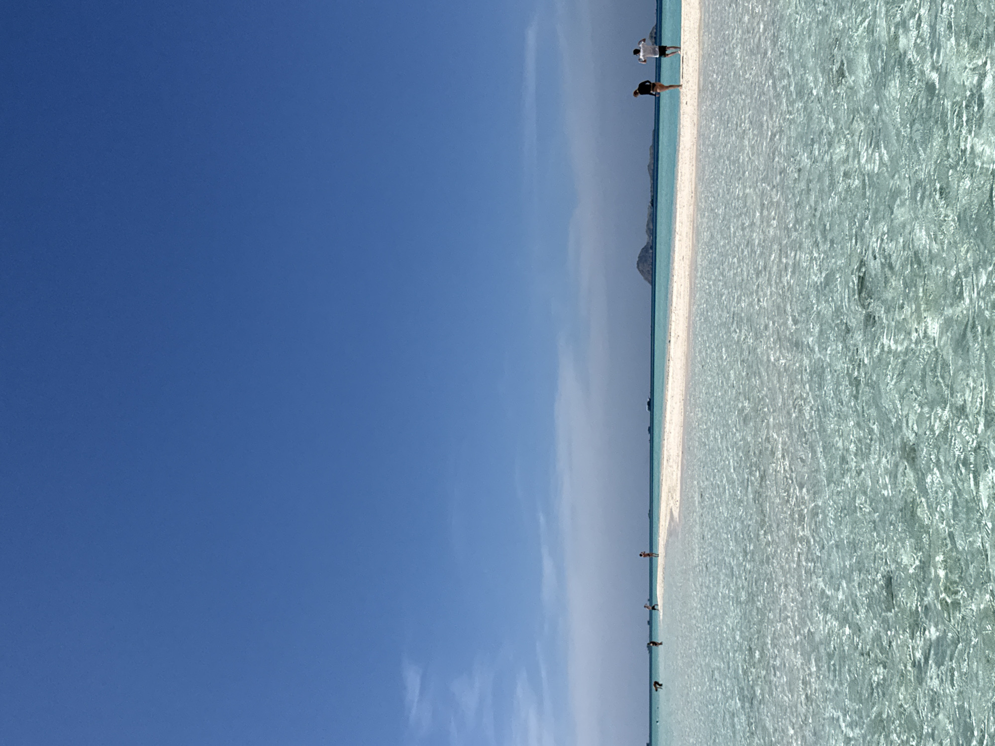 The image depicts a coastal scene in Komodo, Indonesia. A narrow strip of white sand beach divides the clear, turquoise water from the blue sky. Several people are seen standing and walking along the shoreline and in the water. A distant landmass is visible on the horizon. The sky is mostly clear with some scattered clouds.