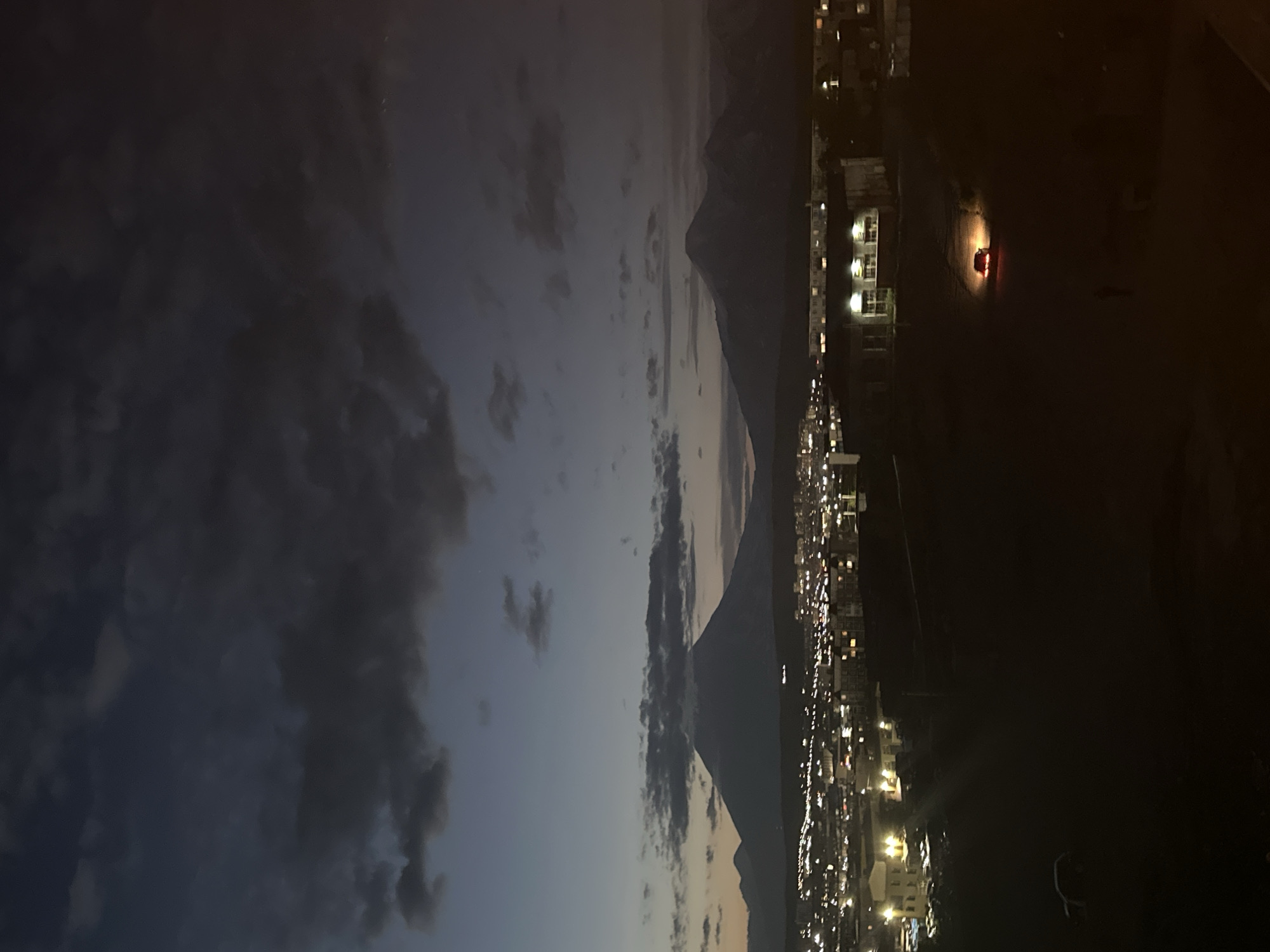 This is a high-angle, long shot photograph of Petropavlovsk-Kamchatsky, Russia, taken during twilight. In the background, two prominent volcanoes dominate the skyline, with the larger, snow-capped volcano in the left half of the frame appearing more distinct. The sky is a deep, dusky blue, streaked with dark, heavy clouds that are beginning to dissipate. Below the volcanoes, the city lights twinkle, creating a soft glow over the urban landscape. Buildings of various sizes and shapes are visible, some appearing to be residential areas and others more commercial or industrial. The foreground is dark and indistinct, appearing to be a sloped terrain, possibly a hill or elevated ground overlooking the city. A single red car is visible in the middle ground, its headlights illuminating the dark ground around it, suggesting it is stationary or moving slowly. The overall atmosphere is serene and peaceful, with the twilight casting a moody yet beautiful ambiance over the scene.
