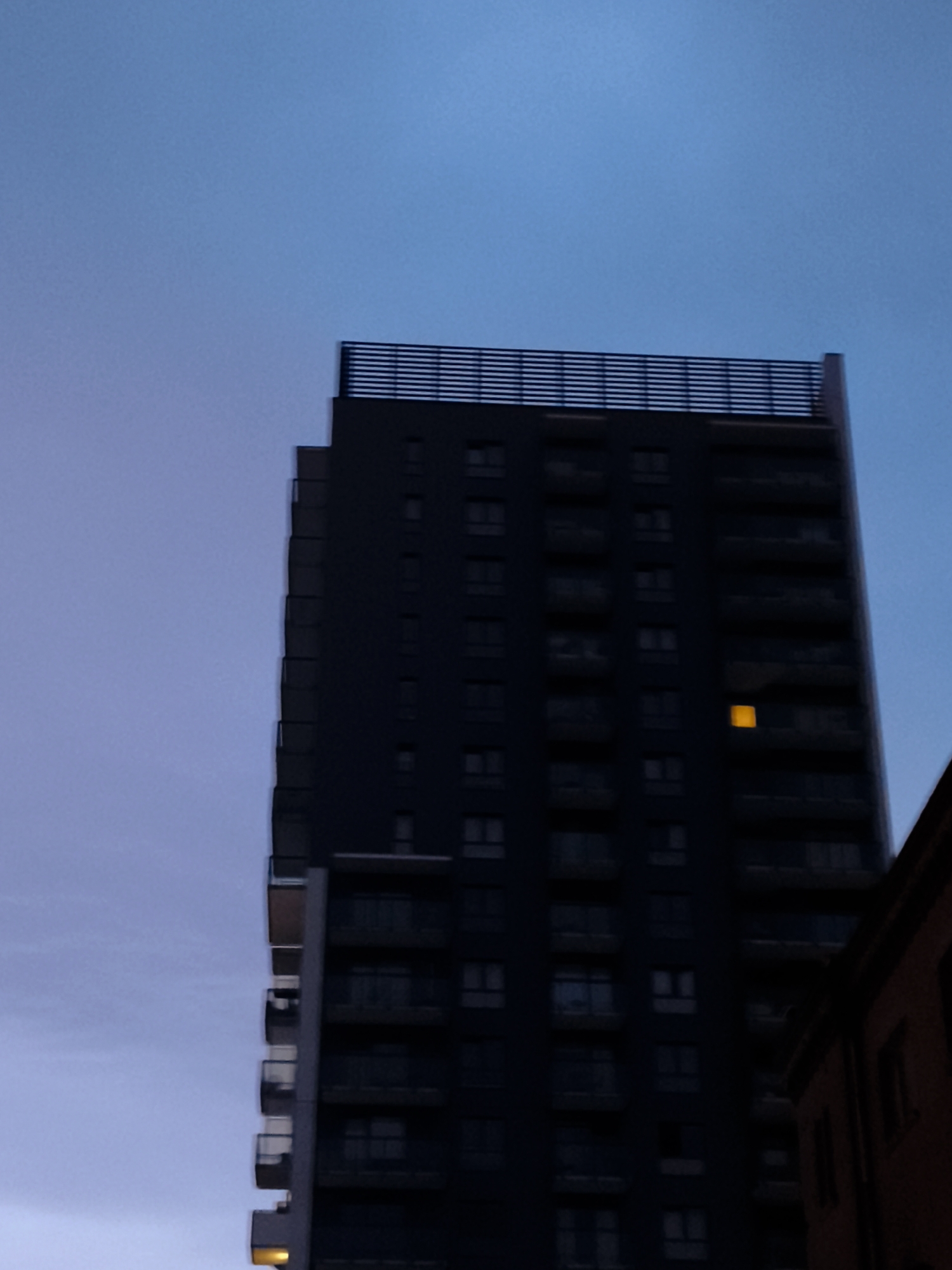 A tall, dark, modern building with many windows and balconies is shown against a twilight sky. The building's architecture is sleek and geometric, with a horizontal railing structure at the top. One window on the lower section of the building is illuminated with a warm, yellow light. To the right, a portion of a shorter, brick building is visible, partially obscuring the taller structure. The sky is a gradient of deep blue at the top, fading to a lighter, purplish-blue towards the horizon. The overall impression is one of urban twilight, with a sense of quietude. This building is likely located in Gdańsk, Poland, given its modern architectural style, which is common in urban developments in Polish cities.