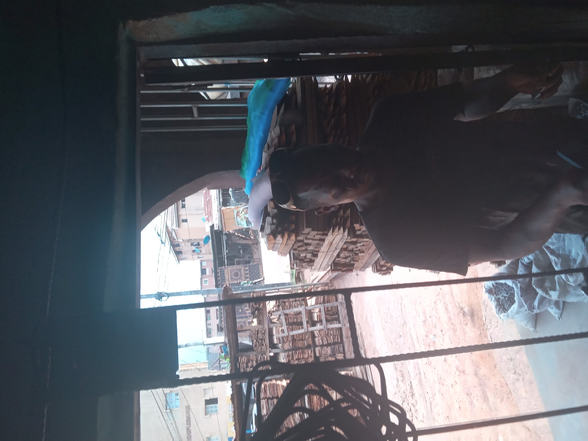 A person is seen wearing a mask, standing in an area with stacks of lumber. Through a metal-barred opening, buildings and streets are visible in what appears to be an urban setting. A bag of what might be nails is also present. The location is identified as Nkpor, Nigeria.