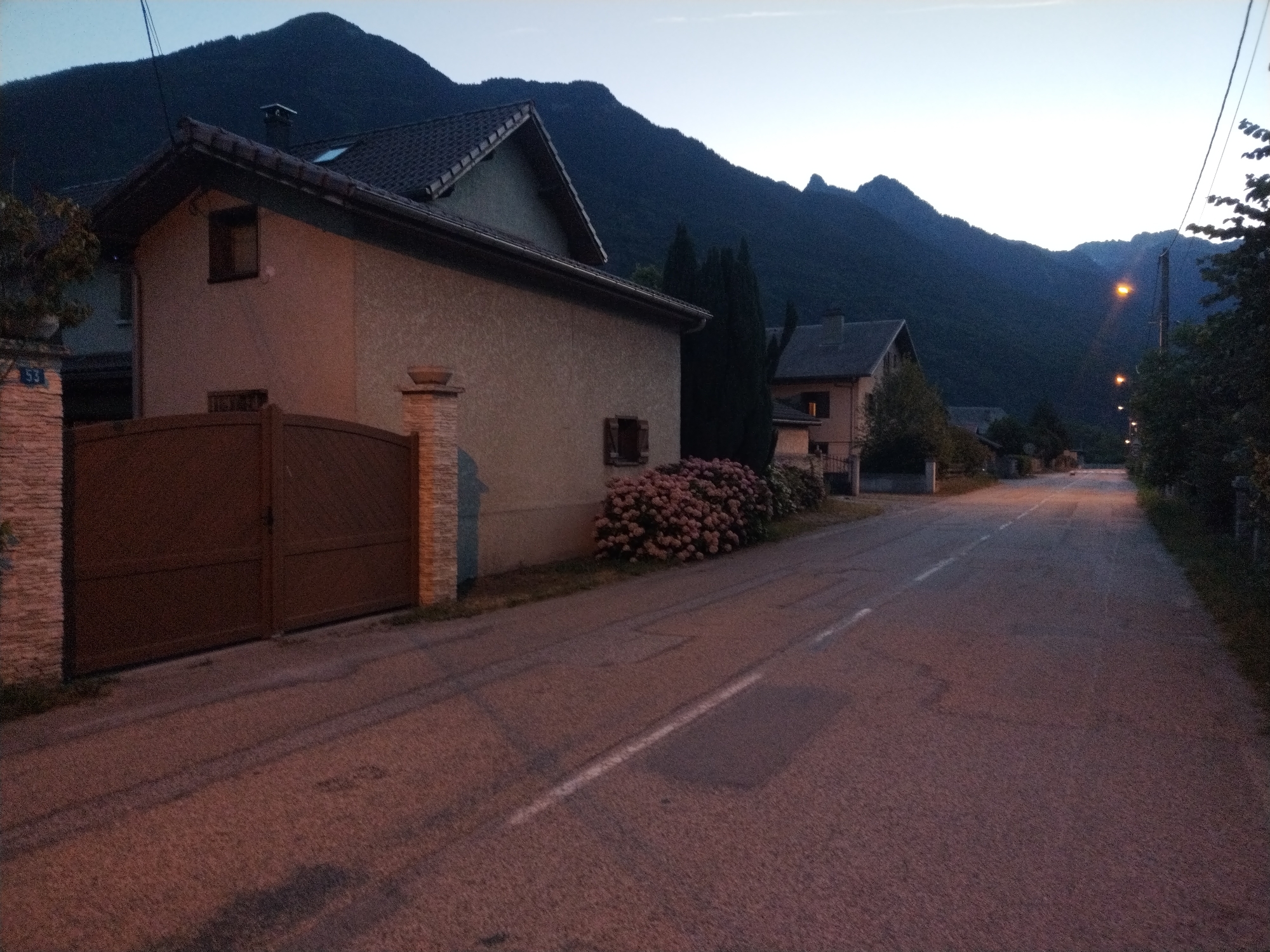 The image depicts a street scene in Saint-Étienne-de-Cuines, France, at dusk. A residential house with a brown gate and a stone pillar is visible on the left. In the background, a dark mountain range dominates the skyline under a pale sky. A road stretches into the distance, lined with streetlights and trees. The overall atmosphere is serene and quiet, suggesting the end of the day.