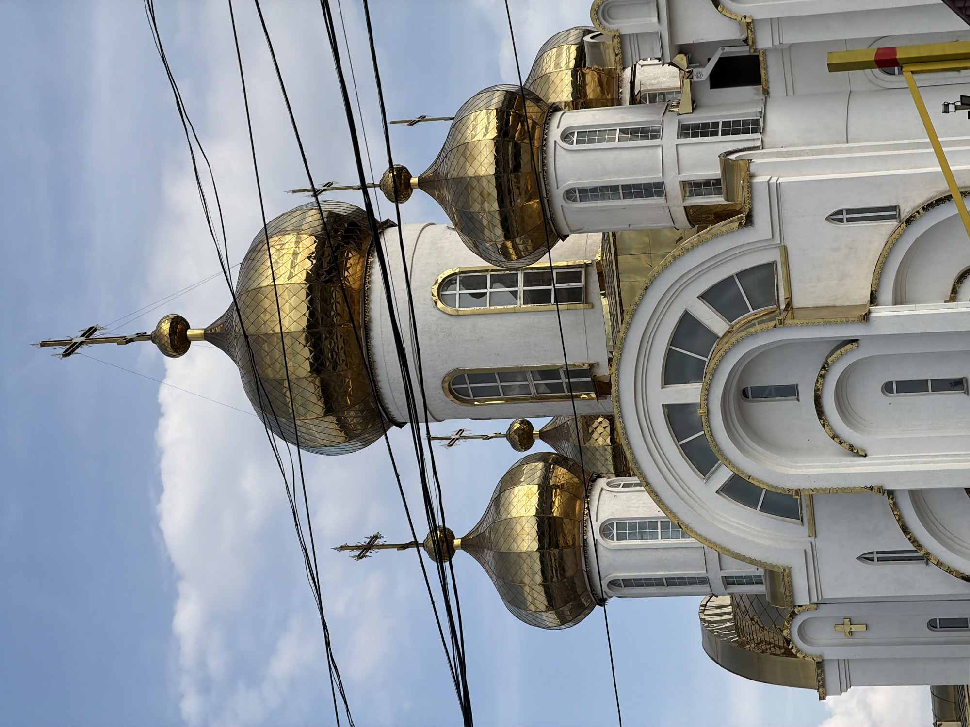 A low-angle, medium shot shows the upper section of a white Orthodox church with golden domes and crosses, set against a partly cloudy blue sky. Multiple black electrical wires crisscross the frame horizontally in the foreground, obscuring parts of the building and sky. The church features several onion-shaped domes, each topped with a cross. The largest central dome is prominently positioned, with smaller domes visible to its sides. The building's facade is white with arched windows, some of which are tall and narrow, while others are large and semicircular. Gold trim accentuates architectural details, including the edges of the domes and around the arched windows. A golden cross is affixed to the lower left portion of the visible facade. In the lower right corner, a portion of a yellow barrier with red accents is visible. The scene appears to be in an urban setting given the presence of electrical wires.