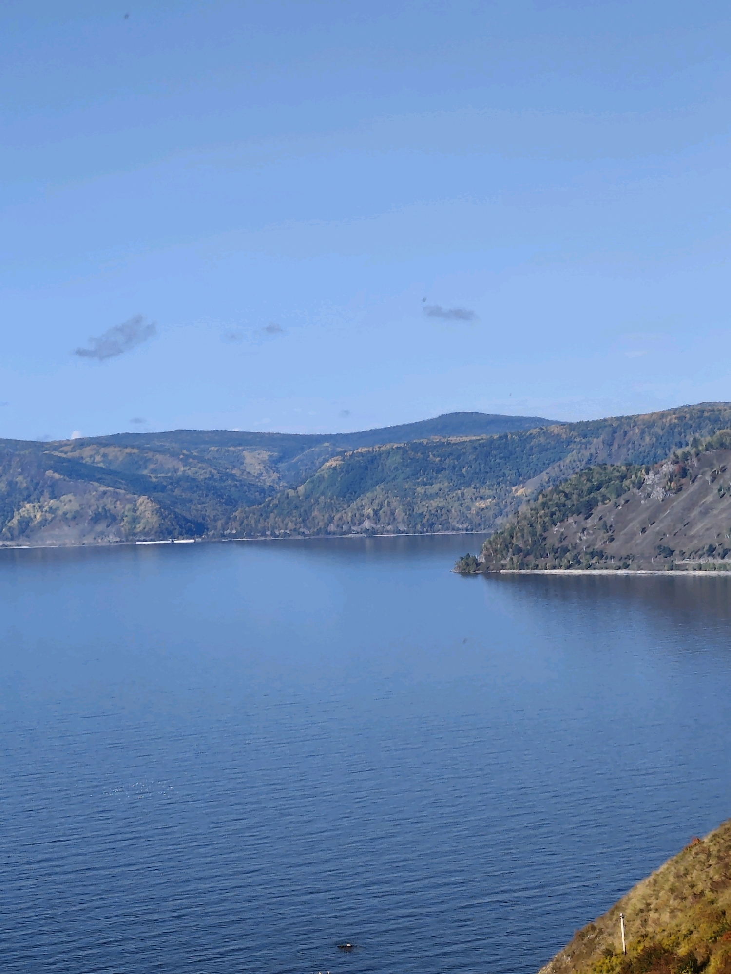 The image depicts a wide, calm body of water with a distant shoreline featuring rolling hills and mountains. The hills are covered with trees displaying autumn foliage, ranging in color from green to yellow and brown. A long, white structure, possibly a barge or a train, is visible on the water near the shoreline. The sky is clear and blue with a few wispy clouds. In the foreground, a steep, grassy bank slopes down towards the water. The overall scene is serene and natural, suggesting a location in a vast, undeveloped area. The location is the city of Baikal, Russia.