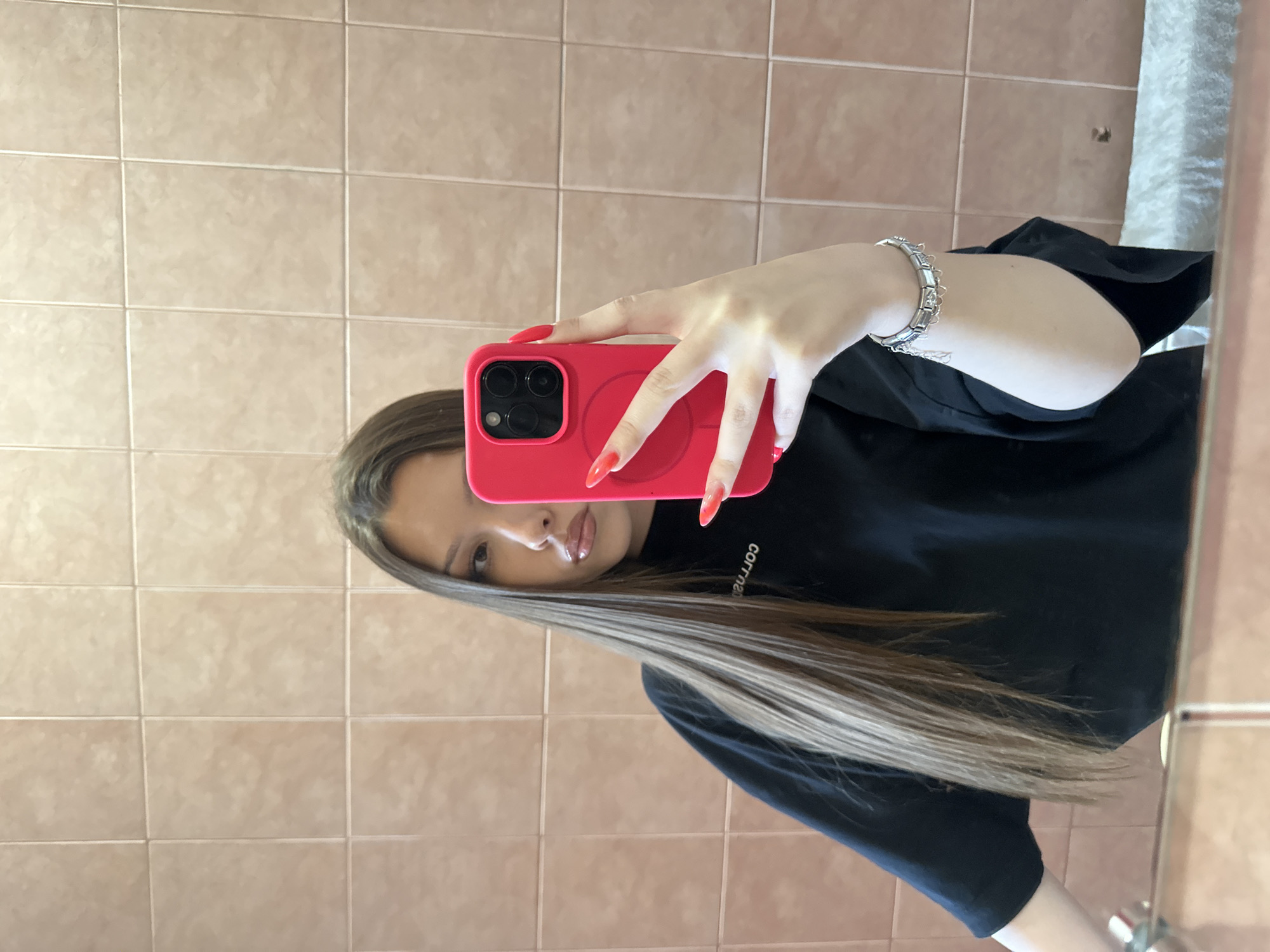A woman is taking a selfie in a bathroom. She is holding a red phone with a black camera module, the screen is pointed towards the mirror. Her nails are long and painted red. She is wearing a black shirt and a silver bracelet on her wrist. Her long brown hair is partially covering her face. The background shows beige tiled walls. This image was taken in the city of Varna, Bulgaria.