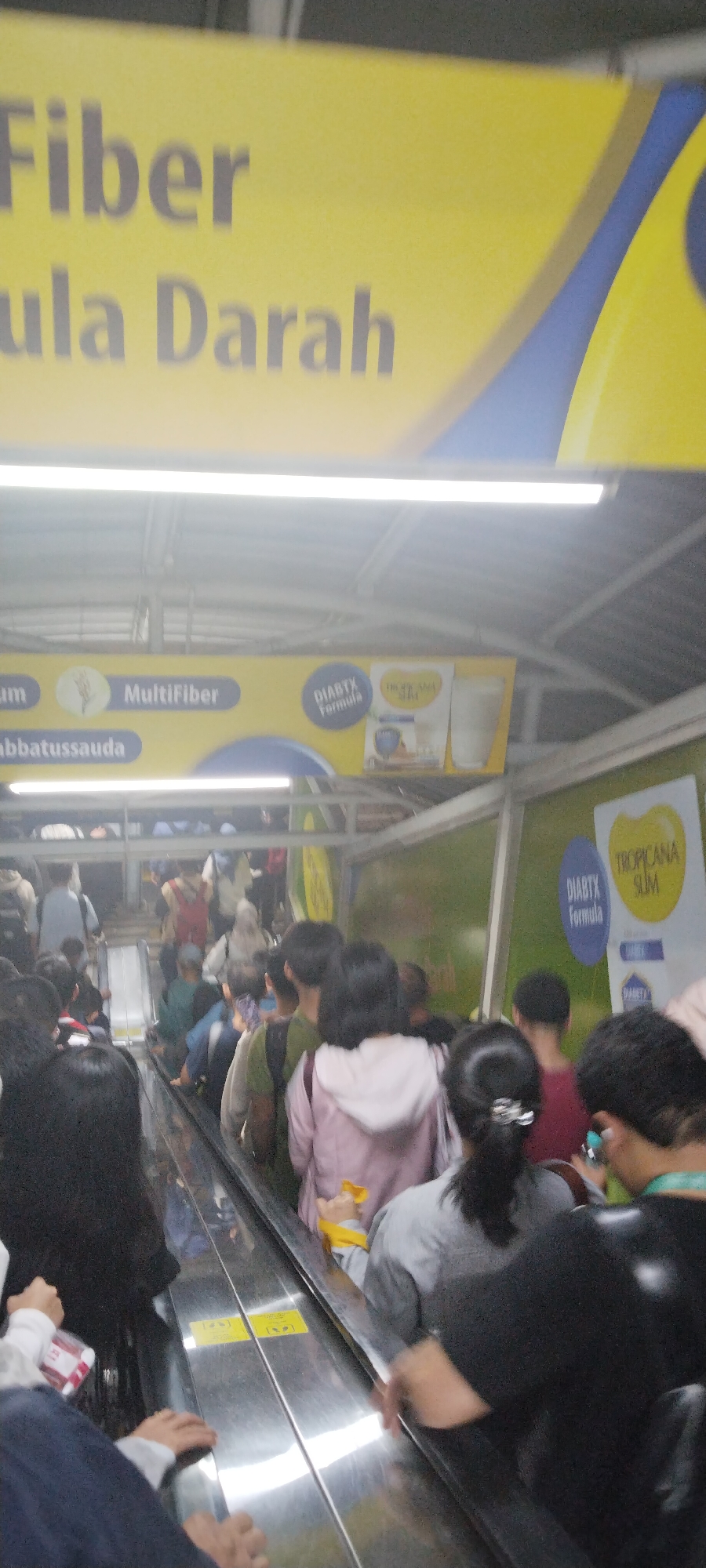 This is a candid, slightly blurred photograph taken from a low angle on an escalator in Central Jakarta, Indonesia. The escalator is stainless steel with yellow warning signs. It appears to be an indoor public space, possibly a mall or transportation hub, with advertisements visible overhead.
The upper part of the frame shows a yellow banner with text in Indonesian, including the words "Fiber" and "Gula Darah" (Blood Sugar). Below this, another yellow banner features the text "MultiFiber" and an image of wheat. Further to the right, a blue circular logo with the word "DIABTX FORMULA" is visible.
The escalator is populated with people of various ages and genders, all facing upwards. Most are looking forward or down, some are holding onto the handrail. The individuals are dressed in casual clothing, including t-shirts, hoodies, and jackets. There is a general sense of movement and busyness. The lighting is bright, likely from overhead fluorescent lights, creating some glare on the escalator's surface. The background is somewhat indistinct but suggests a green wall and more advertisements or signage.