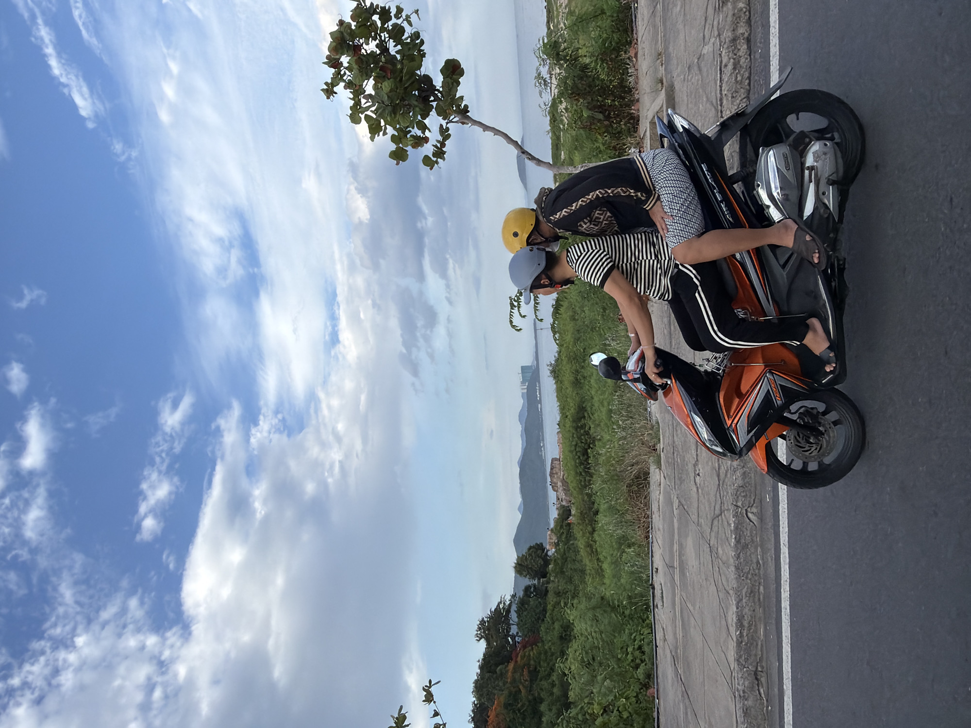 Two individuals are riding on a vibrant orange motorcycle along a paved road. The person in front is wearing a grey helmet and a striped t-shirt with black and white stripes. The person behind, wearing a yellow helmet, has on a black jacket with a gold patterned design and patterned shorts. They are both looking ahead, with the person behind seemingly leaning into the rider. The motorcycle has a silver engine and black accents.
The road is bordered by a concrete sidewalk with visible cracks, and beyond that, a lush green landscape with various trees and shrubs. In the background, a body of water, likely the sea, stretches towards a hazy mountainous coastline. A few buildings are visible on the distant shore. The sky above is a dynamic mix of blue with scattered white clouds, suggesting it might be late afternoon or early evening. This scene is consistent with the coastal areas of Nha Trang, Vietnam, known for its scenic drives along the waterfront.