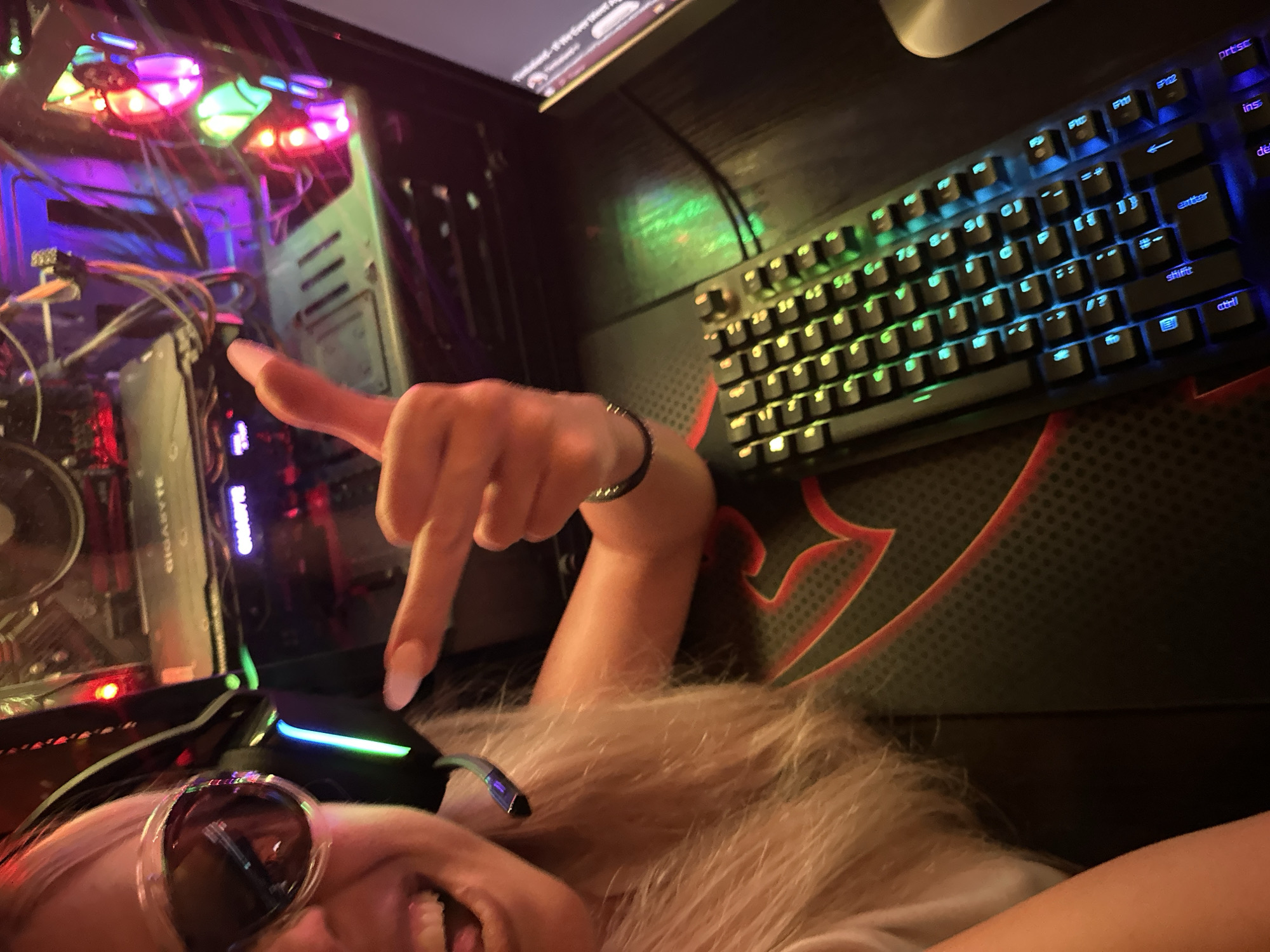 A close-up, high-angle shot shows a blonde woman with light-colored hair and large sunglasses, wearing a white t-shirt, holding up her middle finger in a gesture of defiance. Her mouth is open, and her tongue is slightly visible. She appears to be wearing headphones with green and blue LED lights. Next to her, a gaming computer tower is visible, with colorful RGB lights illuminating the internal components. A black gaming keyboard with RGB backlighting is in the foreground, on top of a mousepad with a red and black design. The scene is indoors, likely in a room dedicated to gaming, characterized by the presence of the computer, keyboard, and headphones. The overall lighting is dim, with the RGB lights of the computer and keyboard providing the primary illumination. There are no discernible landmarks or contextual clues within the image to indicate the specific location in Pazardzhik, Bulgaria.