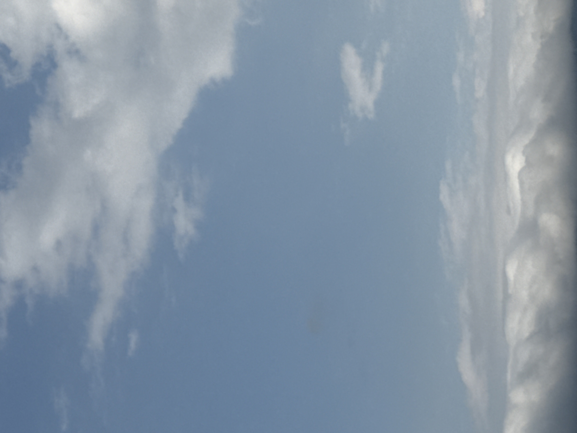 The image is a low-angle shot of a sky filled with clouds. The sky is a clear blue in the upper portion, transitioning to a hazy, overcast sky towards the bottom. Wispy white clouds are scattered across the blue sky, with larger, denser formations of white and gray clouds dominating the lower part of the frame. The lighting suggests it is daytime, possibly late afternoon or early evening, given the diffused light. There are no people, structures, or identifiable landmarks visible in the image. The scene appears to be an open sky, devoid of any specific urban or natural context.