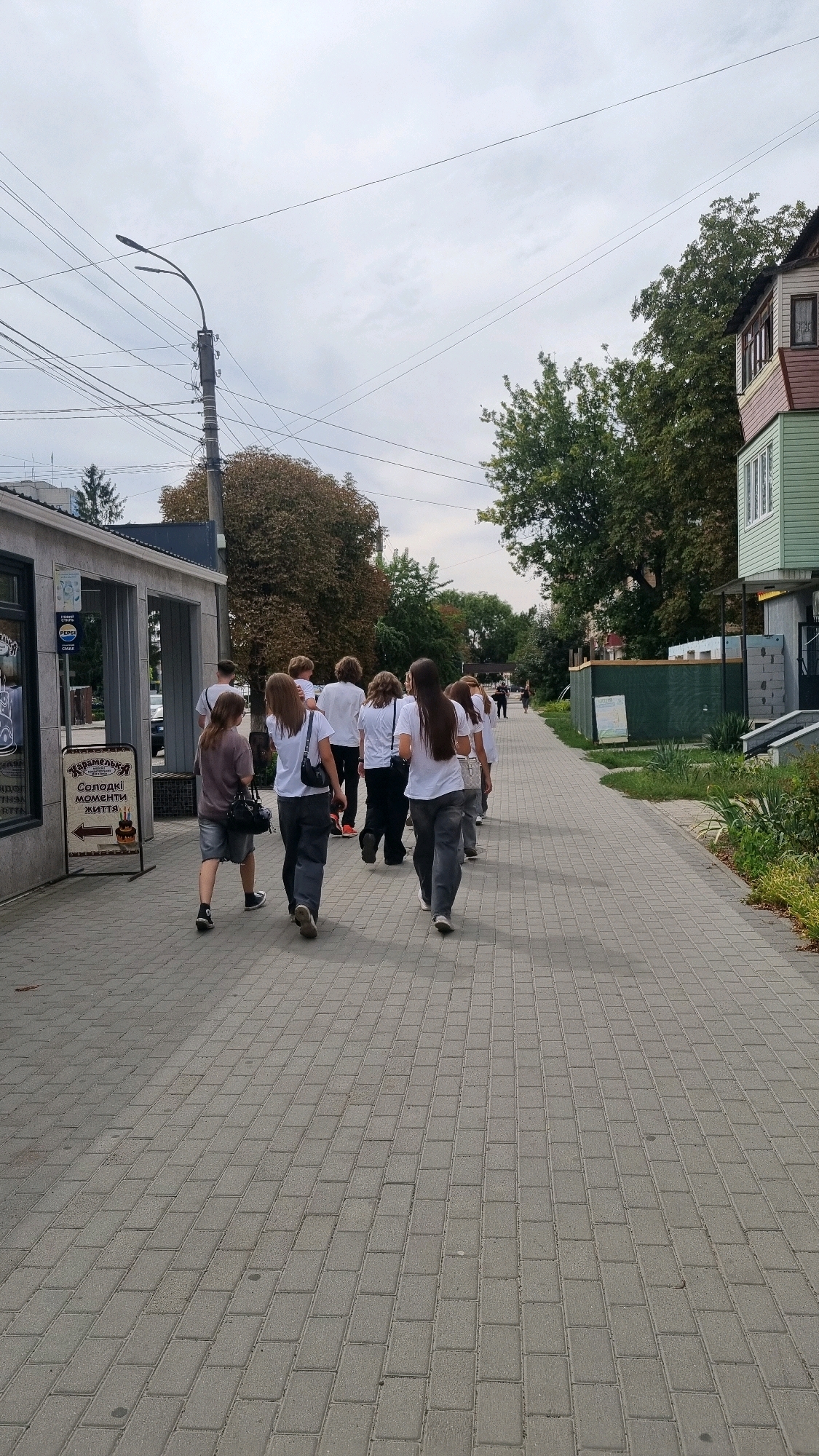 A group of approximately ten young people, predominantly female, wearing predominantly white shirts and dark pants or shorts, walk away from the camera along a paved sidewalk. The sidewalk is constructed of rectangular gray pavers laid in a running bond pattern. The scene is outdoors, with buildings and trees lining the sidewalk. On the left, a modern building with a stone facade and large windows is visible, featuring a prominent sign with Ukrainian text and an image of a cake, indicating a confectionery or bakery. A streetlamp with multiple wires extends from a pole on the left side of the image. On the right, a multi-story building with a green facade and dark trim is situated closer to the sidewalk, with more trees and vegetation in the background. The sky is overcast and gray. The overall impression is a casual daytime scene on a city street in Smila, Ukraine.