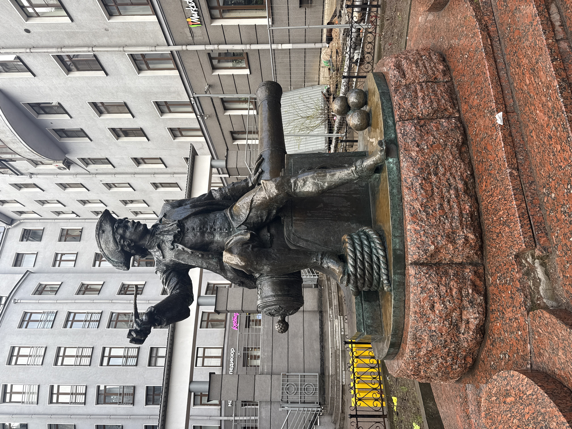 The image captures a bronze statue depicting a man in historical naval attire, complete with a hat, coat, and holding a lit torch. The statue appears to be sitting near a canon with cannonballs in the background. The statue is set upon a red granite base with several tiers. Behind the statue, there is a multi-story building with numerous windows. A sign with "Market" written in a colorful font is visible on the building. A fence is visible in the background. The photograph appears to have been taken in the city of Saint Petersburg, Russia.