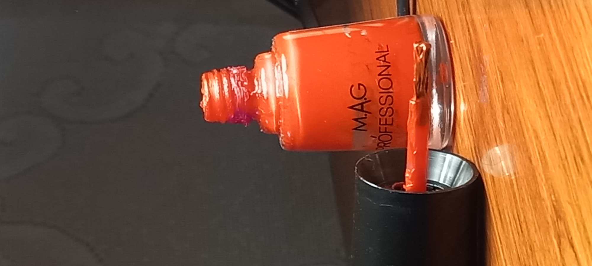 An eye-level shot captures an open bottle of orange nail polish lying horizontally on a surface, likely in Kryvyi Rih, Ukraine. The clear glass bottle contains a vibrant, almost coral-colored nail polish. The brand "M.A.G PROFESSIONAL" is clearly visible in black text on the bottle's side.
The screw threads at the neck of the bottle show remnants of dried orange nail polish, with a hint of purple discoloration, suggesting prior use. The nail polish brush, saturated with the same orange polish, extends from the bottle and dips into the detached, upright black cap, which appears to be a metallic-lined applicator base.
The scene is set indoors, with the right side of the image dominated by a warm, light brown wooden surface, showing distinct grain. The left side of the image features a dark, seemingly textured or patterned surface. The lighting appears to be ambient indoor light, casting subtle reflections on the bottle and the wooden surface. No people are visible.