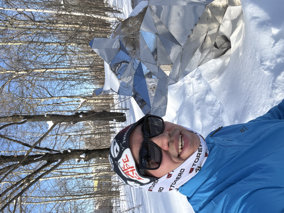 An adult male is depicted in a selfie taken outdoors during winter. He is positioned in the foreground, smiling directly at the camera. He wears black sunglasses, a black and white beanie with "BAPE" and "ЗИМА" visible text, a white neck gaiter patterned with "TORERO," and a bright blue zippered jacket also bearing a "TORERO" logo. To his right, a large, geometrically faceted sculpture of a wolf's head and upper body, constructed from reflective metallic panels, stands partially covered in snow. The sculpture's upper surfaces retain accumulated snow. The environment is characterized by extensive snow cover on the ground and numerous bare deciduous trees against a clear blue sky in the background. Distant architectural elements are vaguely discernible through the tree line. This scene is situated in Victory Park, Kazan, Russia.