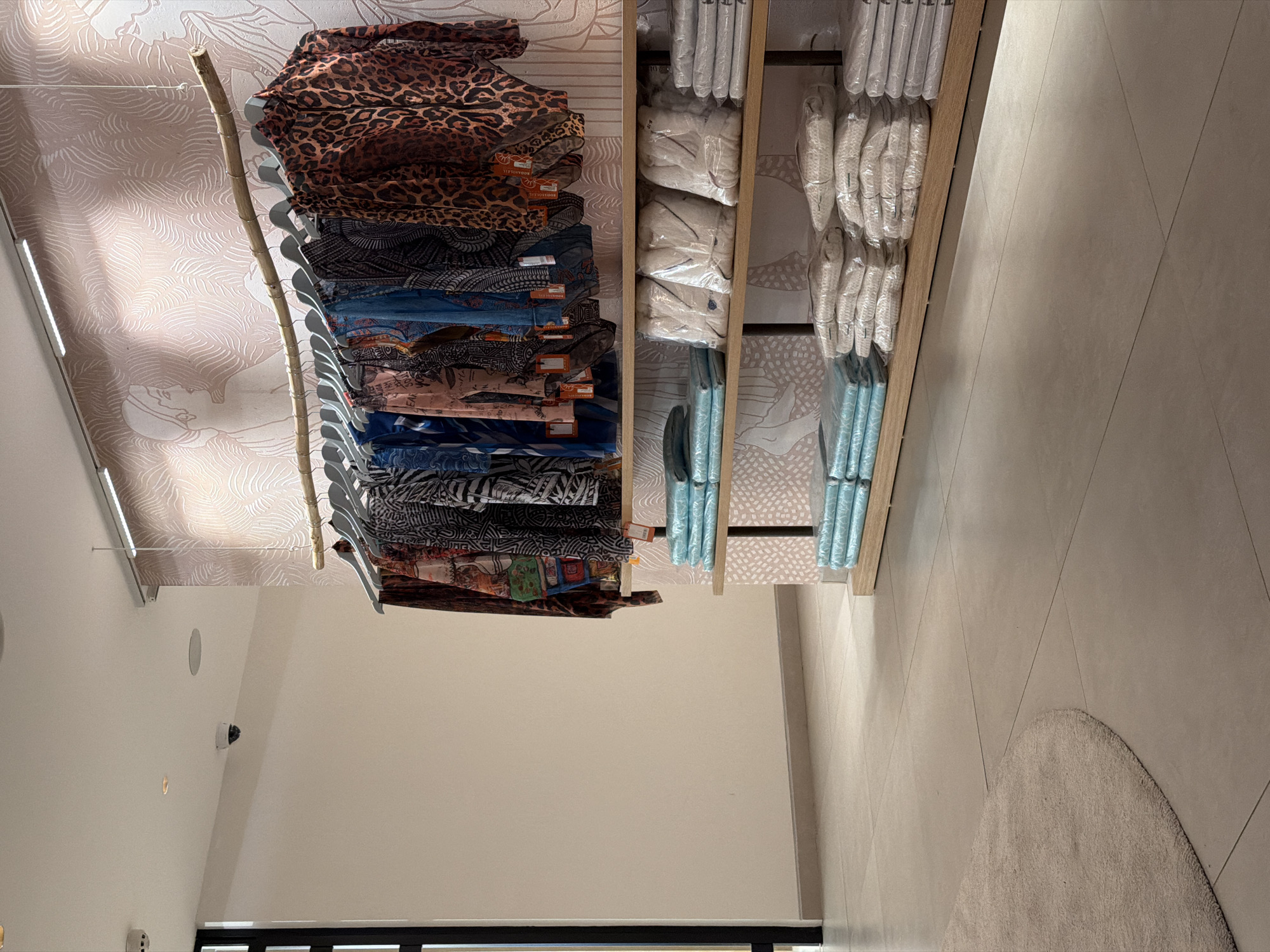 The image depicts a retail store interior, possibly a boutique, featuring a clothing display and shelving. On the right side, a rack of clothing is hung on a natural wood branch. The garments display various patterns, including leopard print and geometric designs. Below the clothing rack, wooden shelves are organized with neatly folded items, some wrapped in plastic. These items appear to be textiles, such as towels or linens, in shades of light blue and white. The background wall is decorated with a light pink wallpaper featuring an abstract pattern and faint outlines of figures. A security camera is visible on the ceiling in the upper left portion of the image. The flooring consists of large, light-colored tiles, with a section of a circular beige rug on the left. The lighting appears to be artificial, with overhead fixtures illuminating the display. The overall scene is clean and organized, suggesting a retail environment focused on presentation. The city of Boreti, Montenegro is not evident in this image.