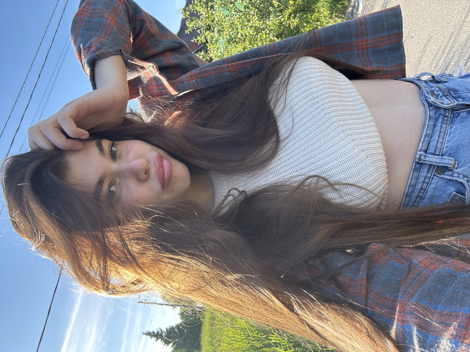 A bright, low-angle outdoor selfie features a young woman with long, dark brown hair, highlighted with golden tones by the sun. She looks directly at the camera with a soft smile and relaxed expression, her left hand gently resting on her head. She is dressed in a light-colored, possibly cream or off-white, ribbed knitted crop top, paired with light blue denim jeans. Over this, she wears an open plaid flannel shirt with a pattern of red, blue, and grey.
The scene is set outdoors under a clear, bright blue sky, indicating sunny weather. In the background, green foliage from trees and bushes is visible, suggesting a natural or semi-urban environment. Faint power lines or cables can be seen against the sky. What appears to be a paved path or road runs along the top right edge of the frame. The strong sunlight and clear sky suggest it is daytime, likely morning or late afternoon. The image is taken in Мещёры, Russia. There is no visible text in the image.