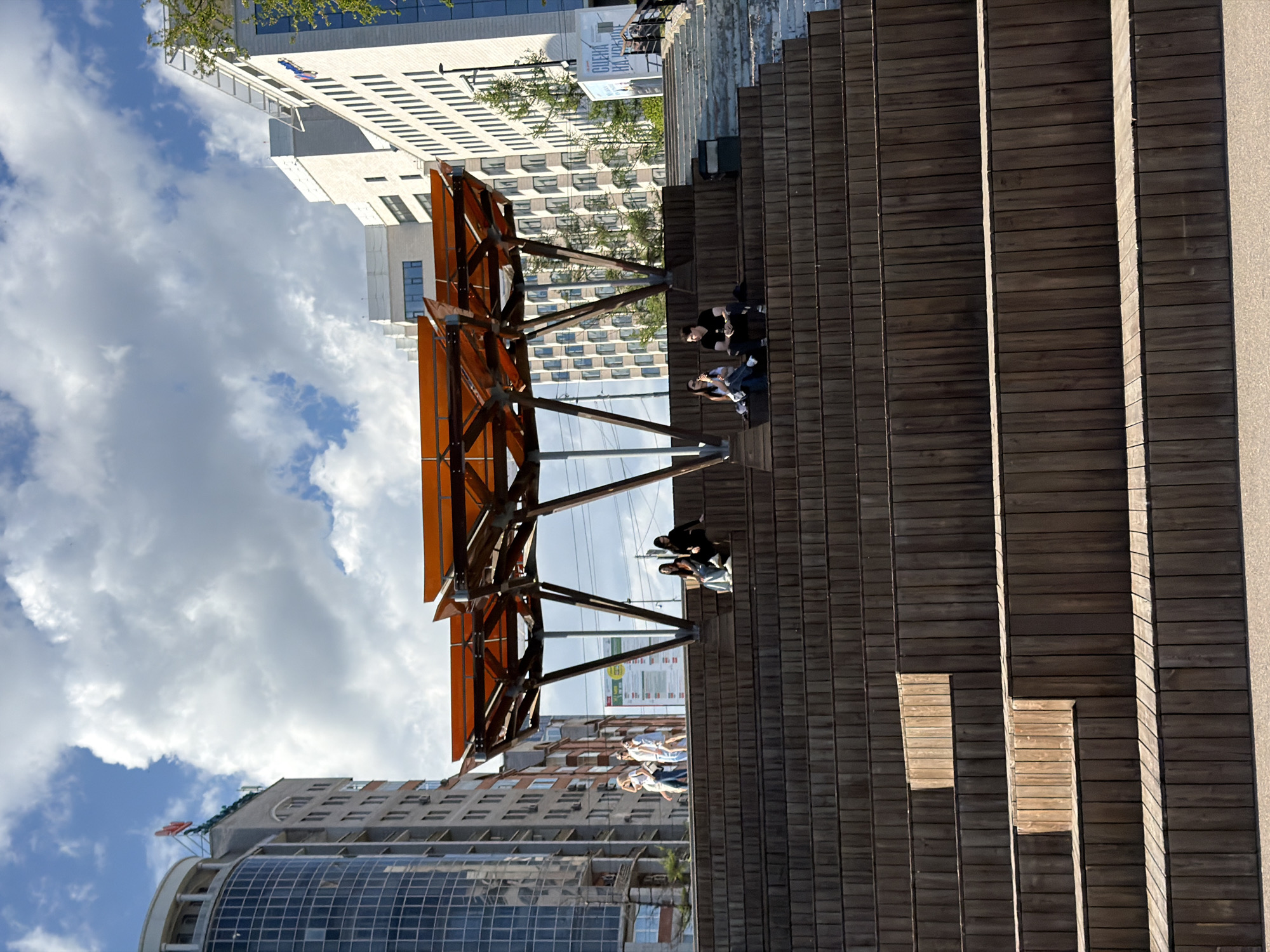 The image shows an outdoor urban space in Krasnoyarsk, Russia. At the center of the frame is a set of stepped wooden platforms, where people are seated and interacting. A modern, geometric shade structure made of metal and wood is above some of the steps. Various buildings are visible in the background, including residential and commercial high-rises. The sky is partly cloudy.