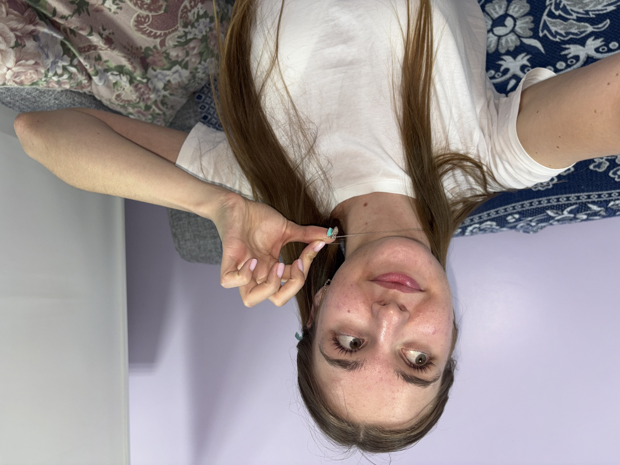 A fair-skinned woman with long brown hair is lying upside down on a bed, holding a turquoise necklace. She is wearing a white t-shirt. The bedding includes a floral-patterned pillow and a blue and white patterned blanket. The background consists of a gray chair and a light purple wall. The location is inferred to be Perm, Russia, based on geographic data not available in the image.