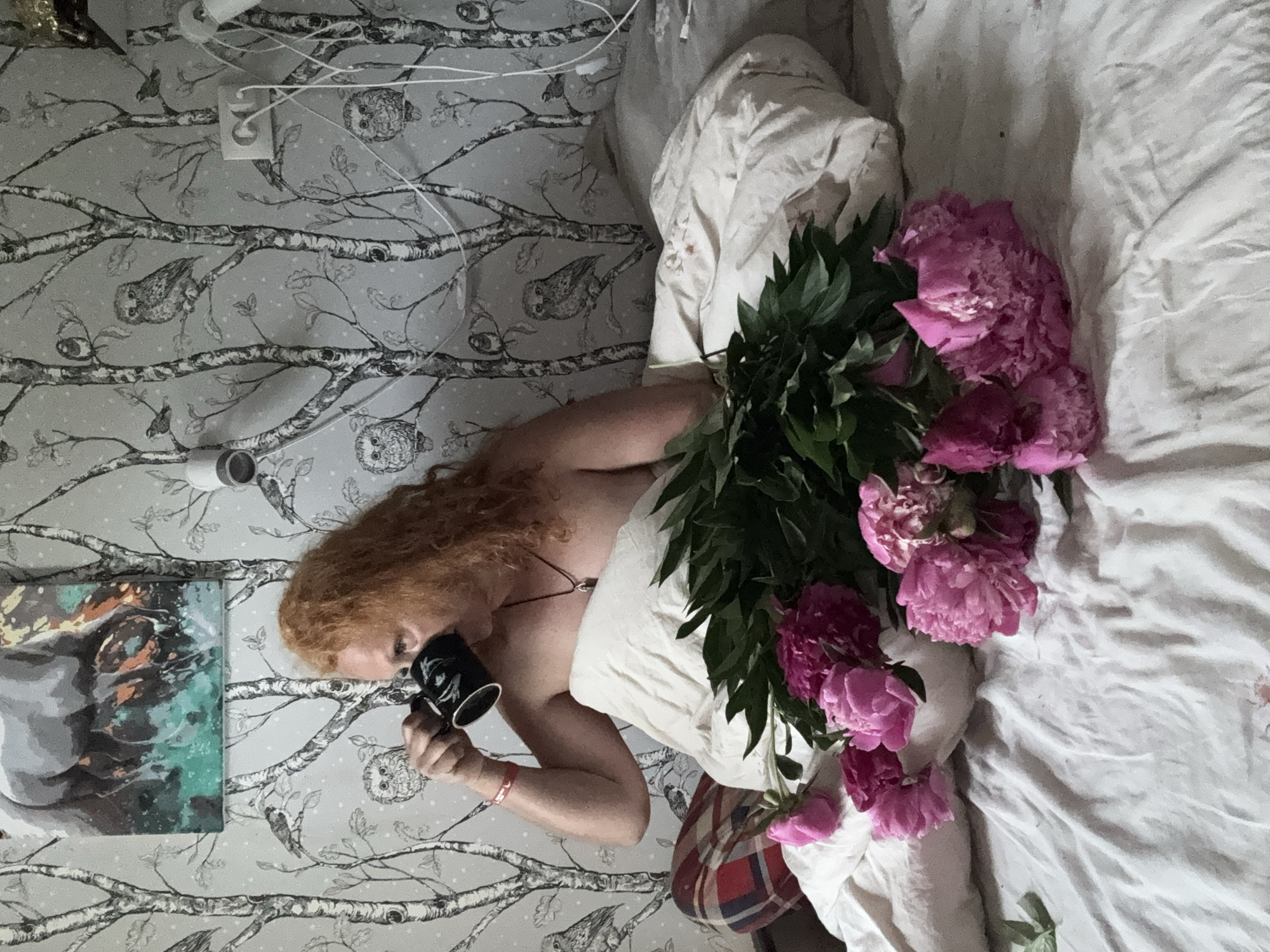 In a bedroom with owl-themed wallpaper, a person with long, red, curly hair is sitting in bed, covered by a white duvet and wearing red and blue plaid pajama bottoms. They are drinking from a black mug with a white design. In front of them is a large bouquet of pink peonies. A painting of a horse is hanging on the wall above the bed. Electrical outlets and cords are visible on the wall. The scene appears to be an indoor domestic setting, likely a bedroom in a private residence. The specific location within the city of Chaikovsky, Russia, cannot be determined from the visual information provided.