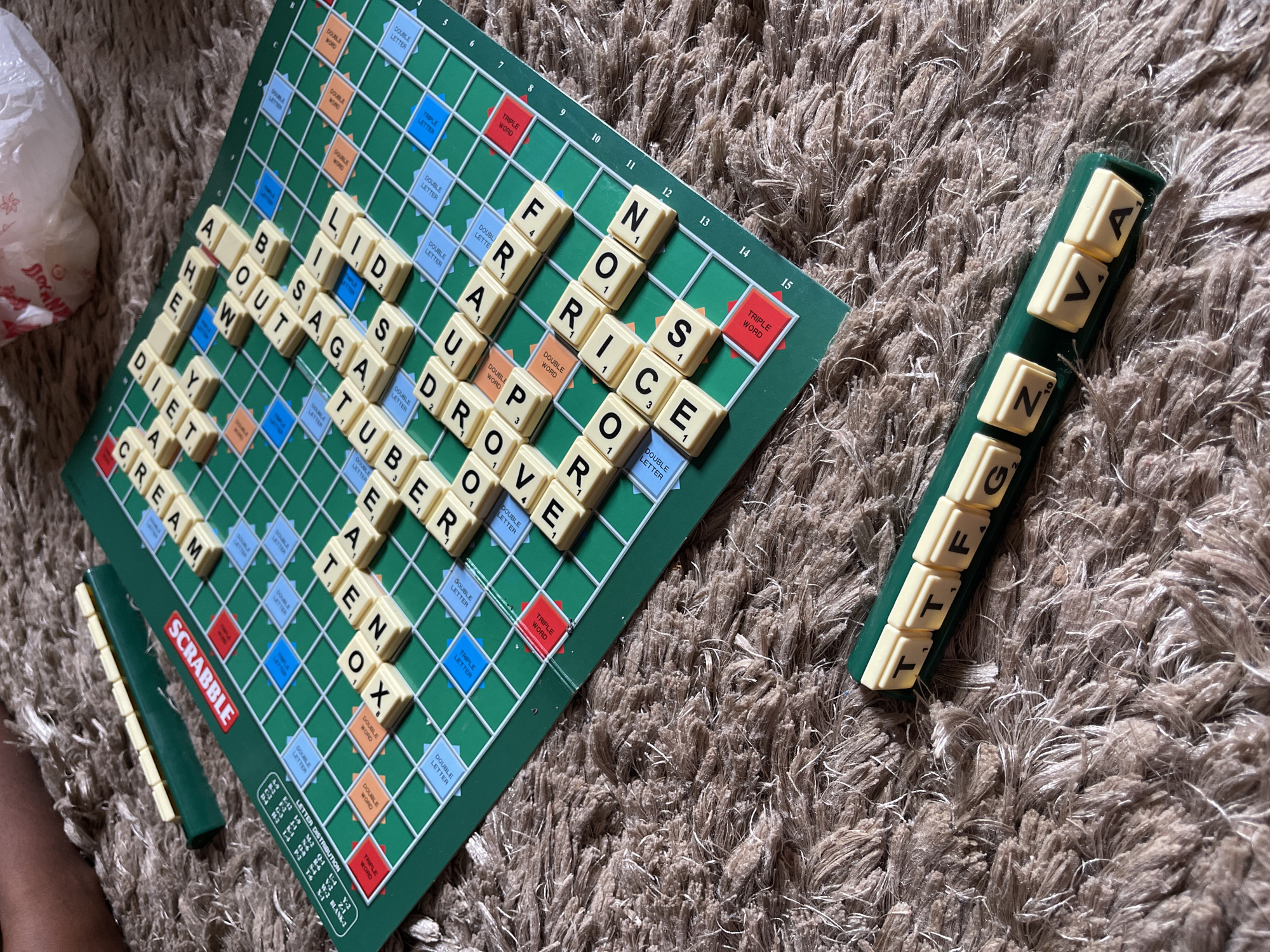 An overhead shot captures a Scrabble board game laid out on a shaggy brown and beige carpet. The green game board is actively in use, with numerous ivory-colored letter tiles arranged to form words. In the upper-left corner, a white plastic bag with a partially visible red logo, reading "DOAN", rests on the carpet. A human hand is visible in the lower-left, near one of the green tile racks.
Words formed on the board include "CREAM," "DIET," "ABOUT," "LIDS," "SAGA," "TUBER," "EATEN," "OX," "DRONE," "PROVE," "FUR," and "NORRICE." Individual letters like "A," "HE," and "W" are also visible. Various squares on the board indicate scoring multipliers such as "DOUBLE WORD," "TRIPLE LETTER," and "DOUBLE LETTER." The "SCRABBLE" logo is prominently displayed on the board's edge. A "LETTER DISTRIBUTION" chart is partially visible in the bottom-left corner of the board.
One green tile rack on the right holds several unplayed tiles, displaying letters "A," "V," "N," "Z," "T," "T," "F," and "G." Another green rack on the left is mostly empty, with some tiles scattered below it. The setting appears to be indoors, likely a home in Kuchingoro, Nigeria, given the provided location context. The lighting suggests ambient indoor conditions.