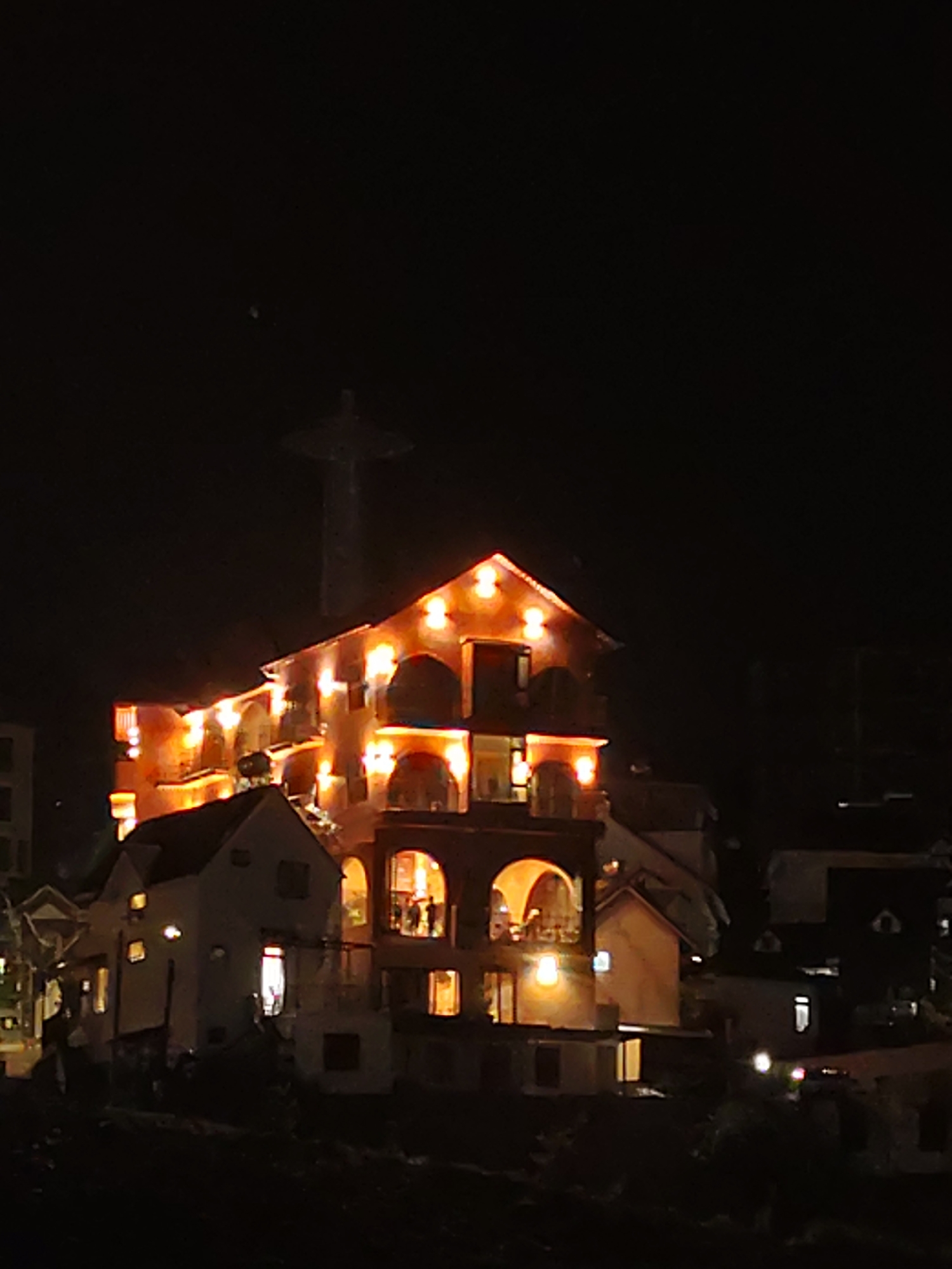 The image shows a night view of Da Lat, Vietnam. A large, ornate building with illuminated arches and lights stands prominently. In the background, a tall structure with a cross on top is visible. Other buildings and lights are scattered around, contributing to the urban landscape. People are visible on one of the balconies in the large building. The overall setting is dark with various points of light.