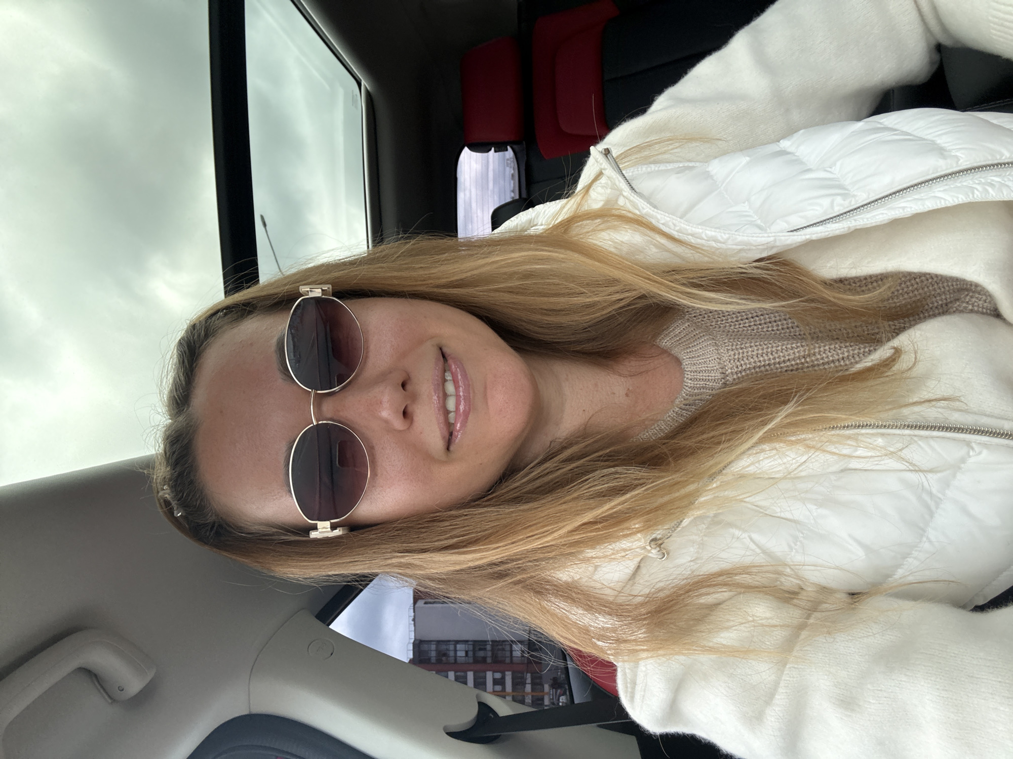 A woman with blonde hair and sunglasses is sitting in the passenger seat of a car, taking a selfie. She is wearing a white puffer vest over a beige sweater. The car has a sunroof, and the sky is cloudy. In the background, a building is visible through the car window. The woman is smiling and looking at the camera.