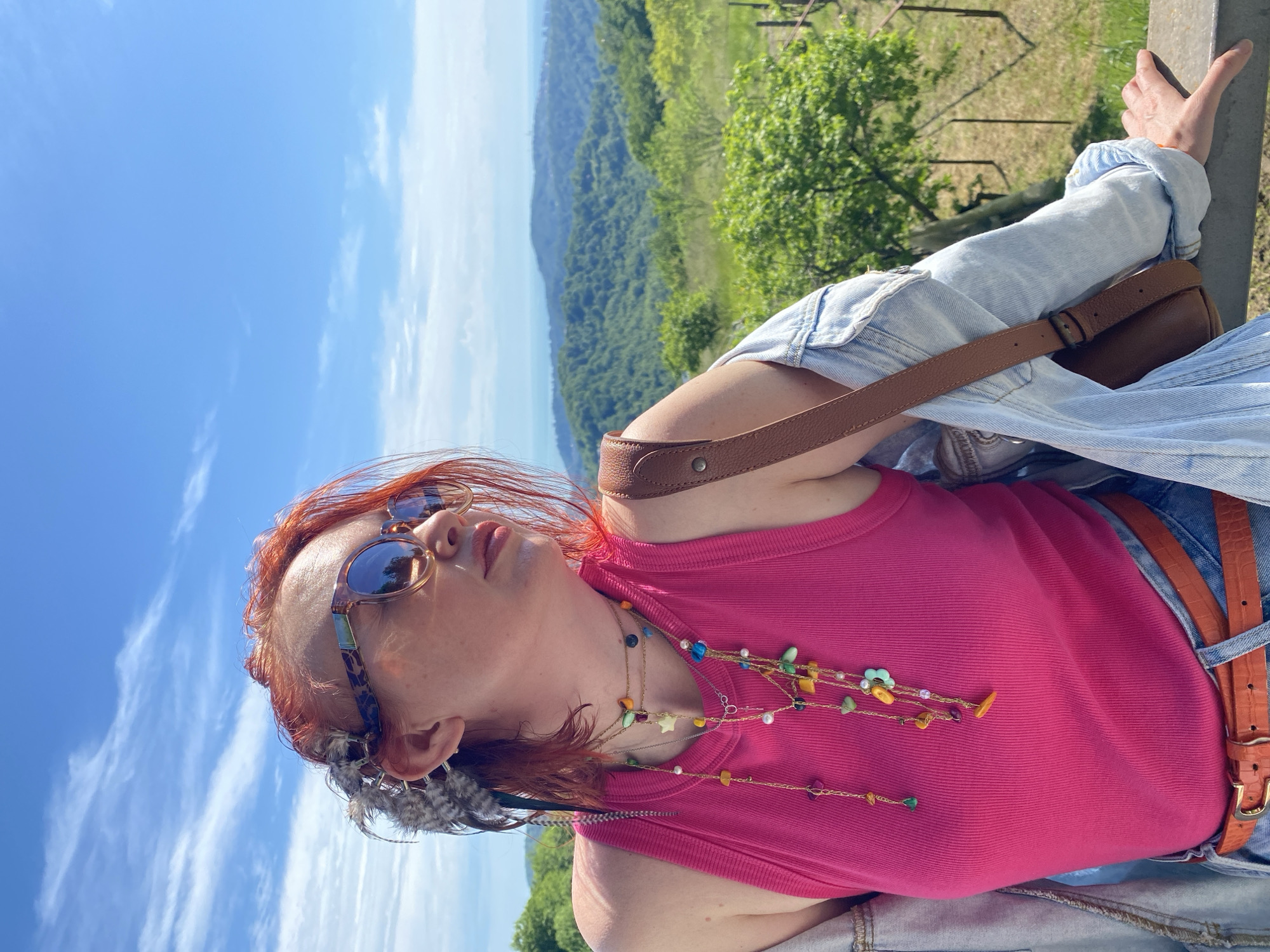 A woman with red hair is outdoors on a sunny day, positioned against a railing with a green landscape in the background and a blue sky. She wears sunglasses, a pink tank top, and a light blue denim jacket. A brown leather bag is slung across her body. The location is not the city of Прогресс, Russia, as it is an outdoor shot.
