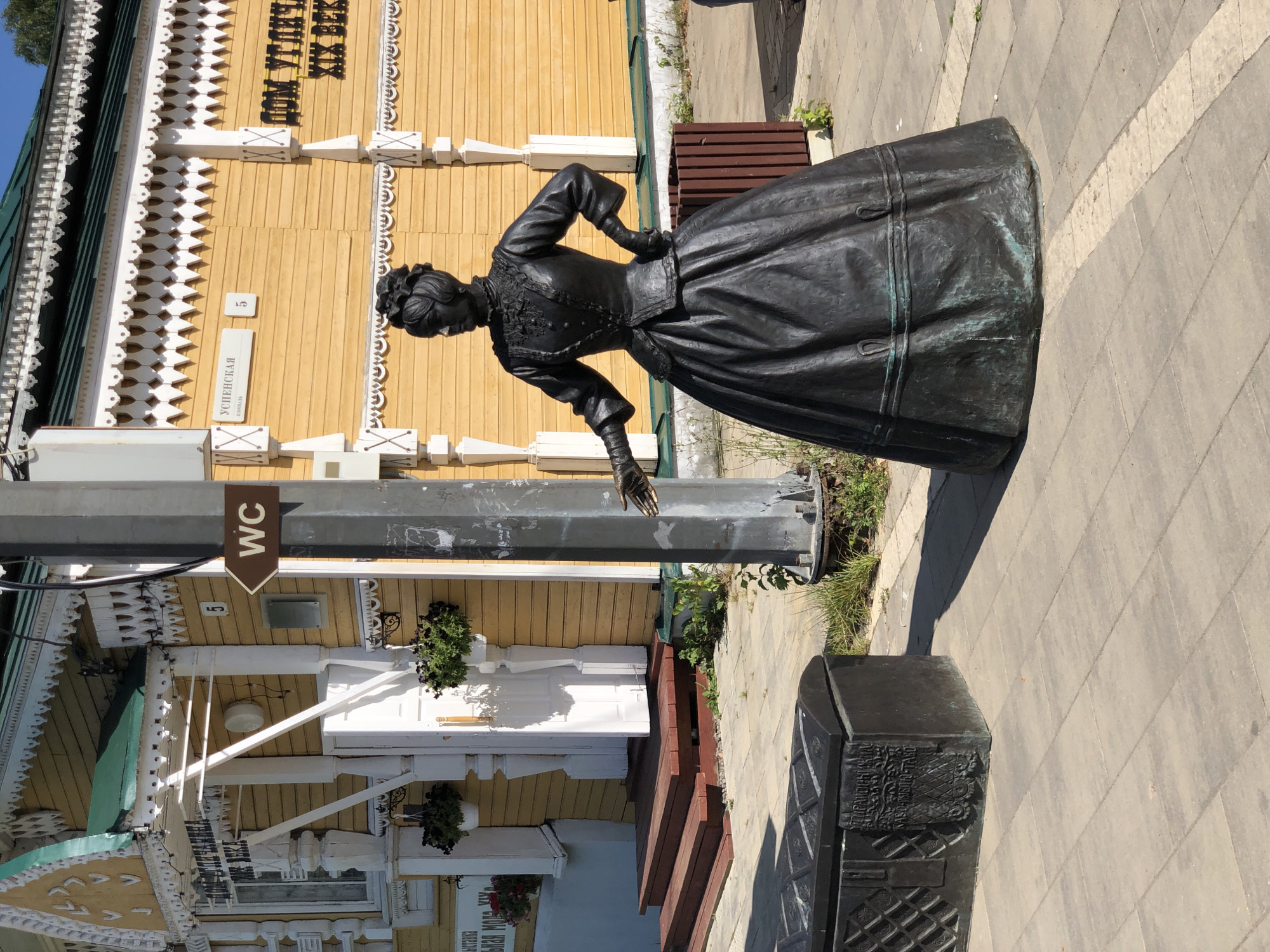 The image shows a bronze statue of a woman in a formal dress, standing on a paved walkway in front of a yellow wooden building. The building has intricate white wooden decorations and signage in Russian. A sign with the letters "WC" and an arrow points to the right, indicating a restroom. To the left of the statue is a dark metal chest with inscriptions and decorative patterns. The scene appears to be in a public area with some greenery and a trash receptacle visible. The statue is positioned to the right of a metal pole. The overall setting suggests a historical or cultural site, likely in Vasil'ki, Russia, given the context provided.