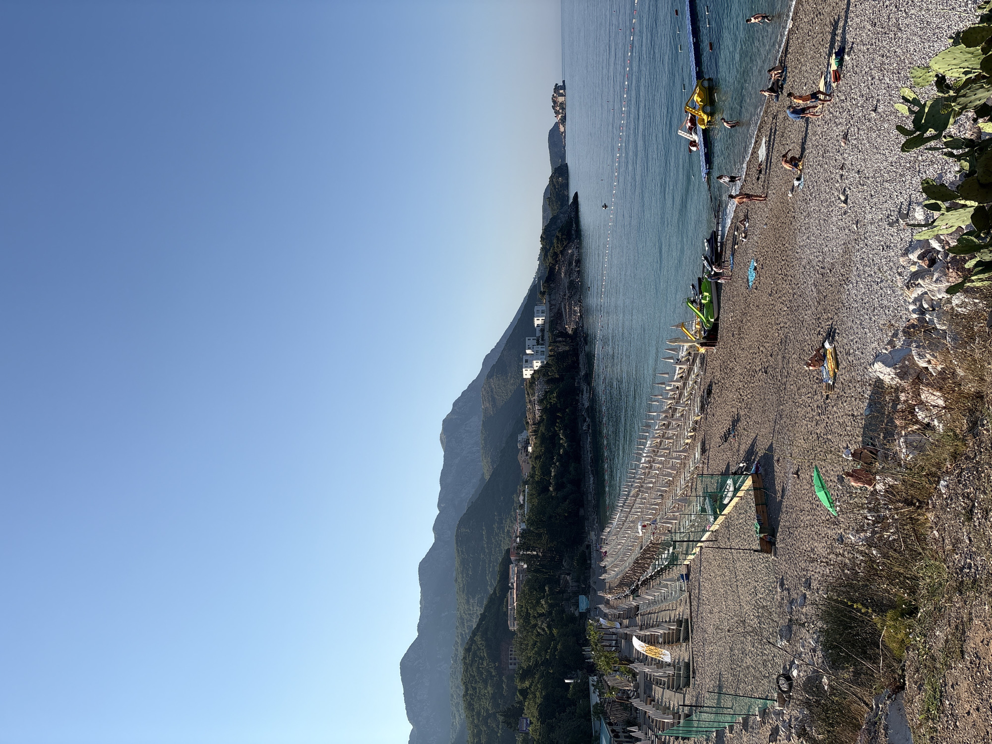 This image depicts a sunny day at Kamenovo beach in Kamenovo, Montenegro. The foreground shows a rocky and sandy shoreline with sparse vegetation. A green umbrella is open with two people sitting beneath it. Beyond them, the beach is densely populated with rows of white lounge chairs and umbrellas, indicating a well-developed resort area. Further down the beach, people are scattered, some sunbathing on towels and chairs, while others are interacting near the water. Several watercraft, including pedal boats and jet skis, are visible near a floating platform in the turquoise sea. The sea stretches out to the horizon, meeting a clear, bright blue sky. In the background, lush green mountains rise, dotted with buildings that suggest a coastal town. A small island with a cluster of buildings is visible in the distance on the right side of the frame. The overall scene conveys a tranquil summer atmosphere at a popular tourist destination.