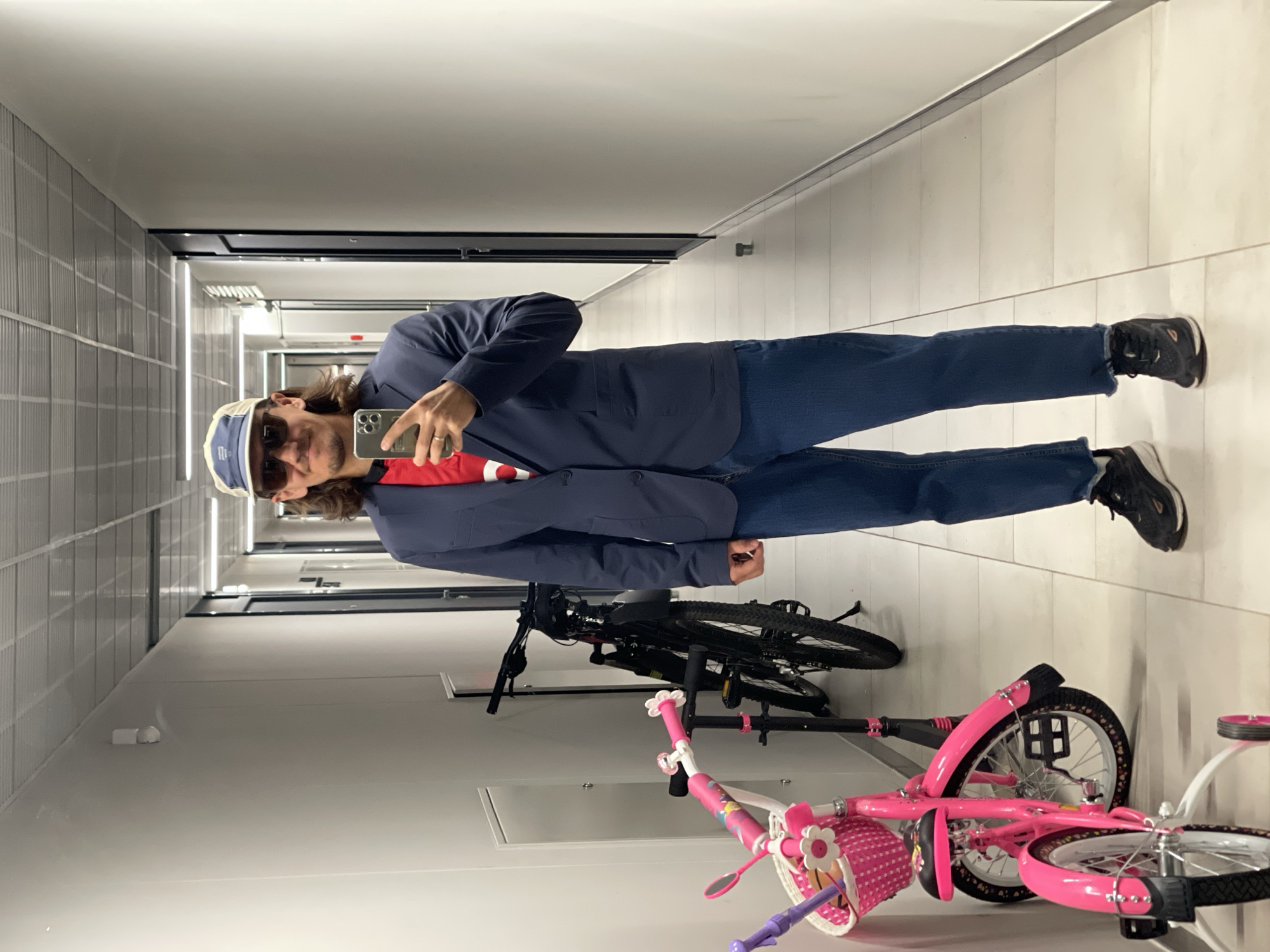 A man in his late 20s or early 30s, with medium-length brown hair, is standing in a brightly lit, white-walled hallway and taking a mirror selfie with his phone. He is wearing a dark blue blazer over a red t-shirt with a white design, blue denim jeans, and black athletic shoes. He also has sunglasses and a light-colored baseball cap on. In the background to his left, there is a pink child's bicycle with training wheels and a basket, and a black bicycle. The hallway has a tiled floor and a drop ceiling with fluorescent lighting. There are doors visible on either side of the hallway in the background. The overall scene appears to be an indoor, communal area.